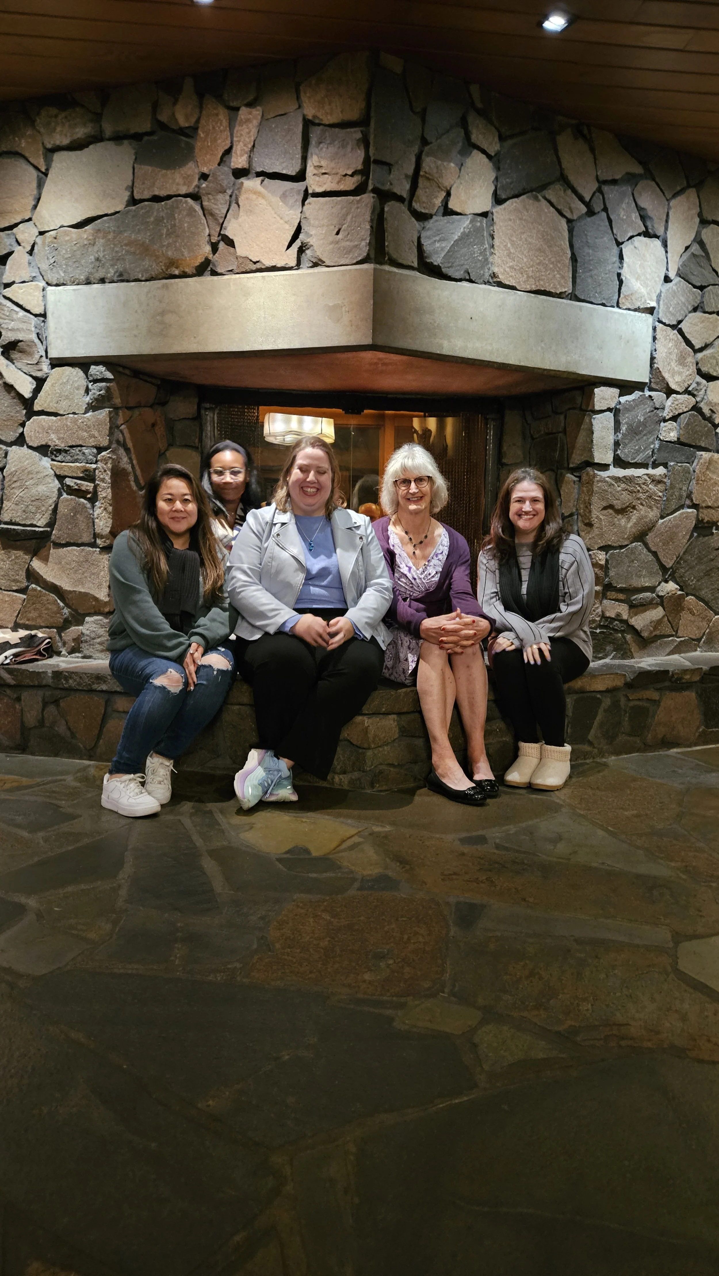 Five women sitting together on a stone bench in front of a stone fireplace, smiling.