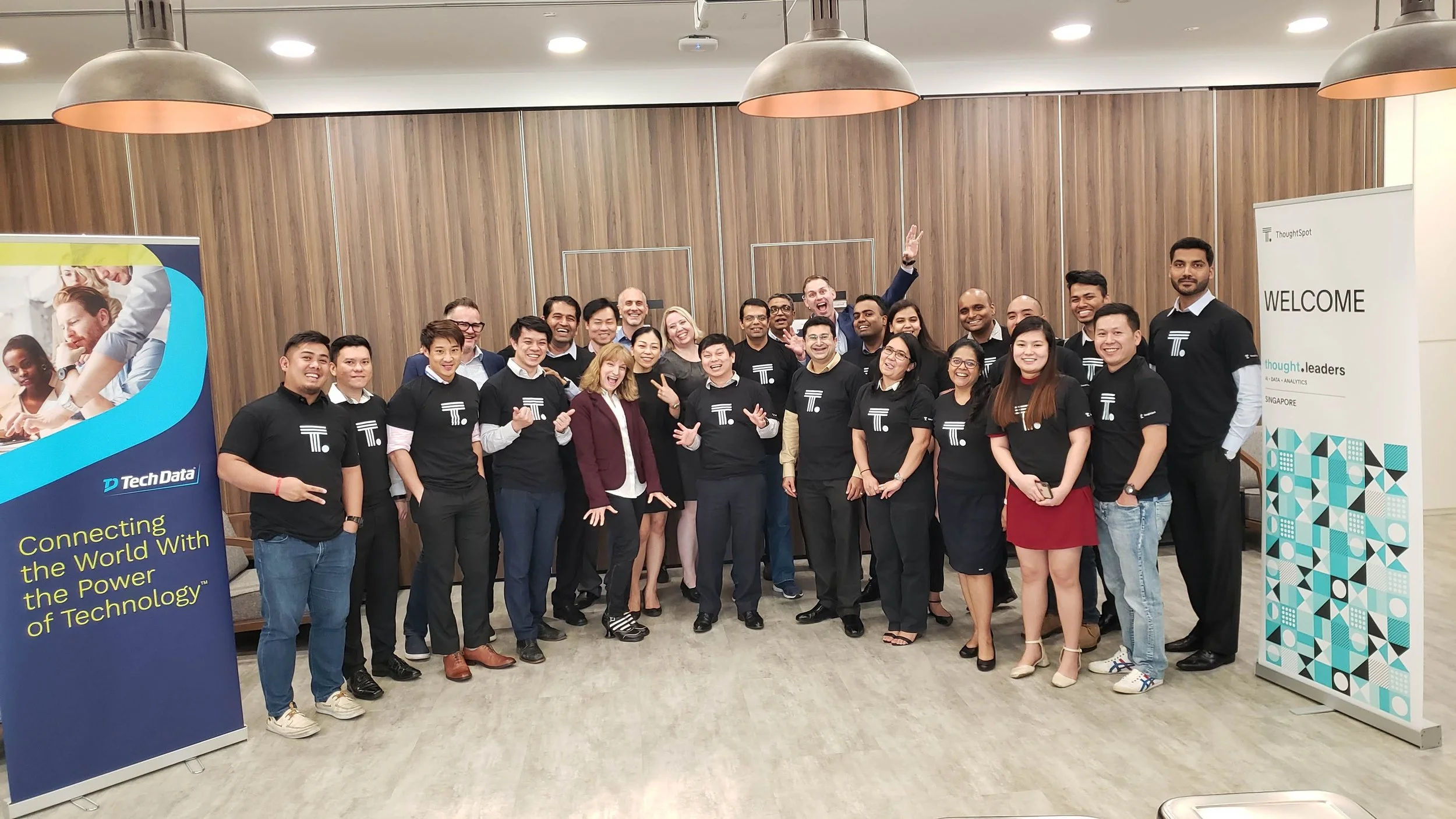 Group of smiling people at a corporate event, standing in a conference room with wooden paneled wall, some wearing black event t-shirts with white logos, and greeting or posing for the photo.