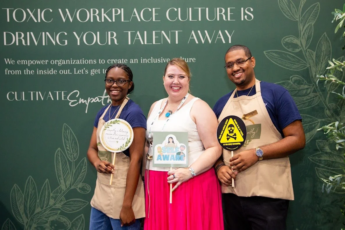 Three people standing in front of a green backdrop with the text 'Toxic Workplace Culture is Driving Your Talent Away' and leaf illustrations. They are smiling, holding signs, and wearing aprons. The woman in the middle wears a white top and pink skirt, and the other two wear navy shirts. The signs include messages about empathy, self-awareness, and safety.