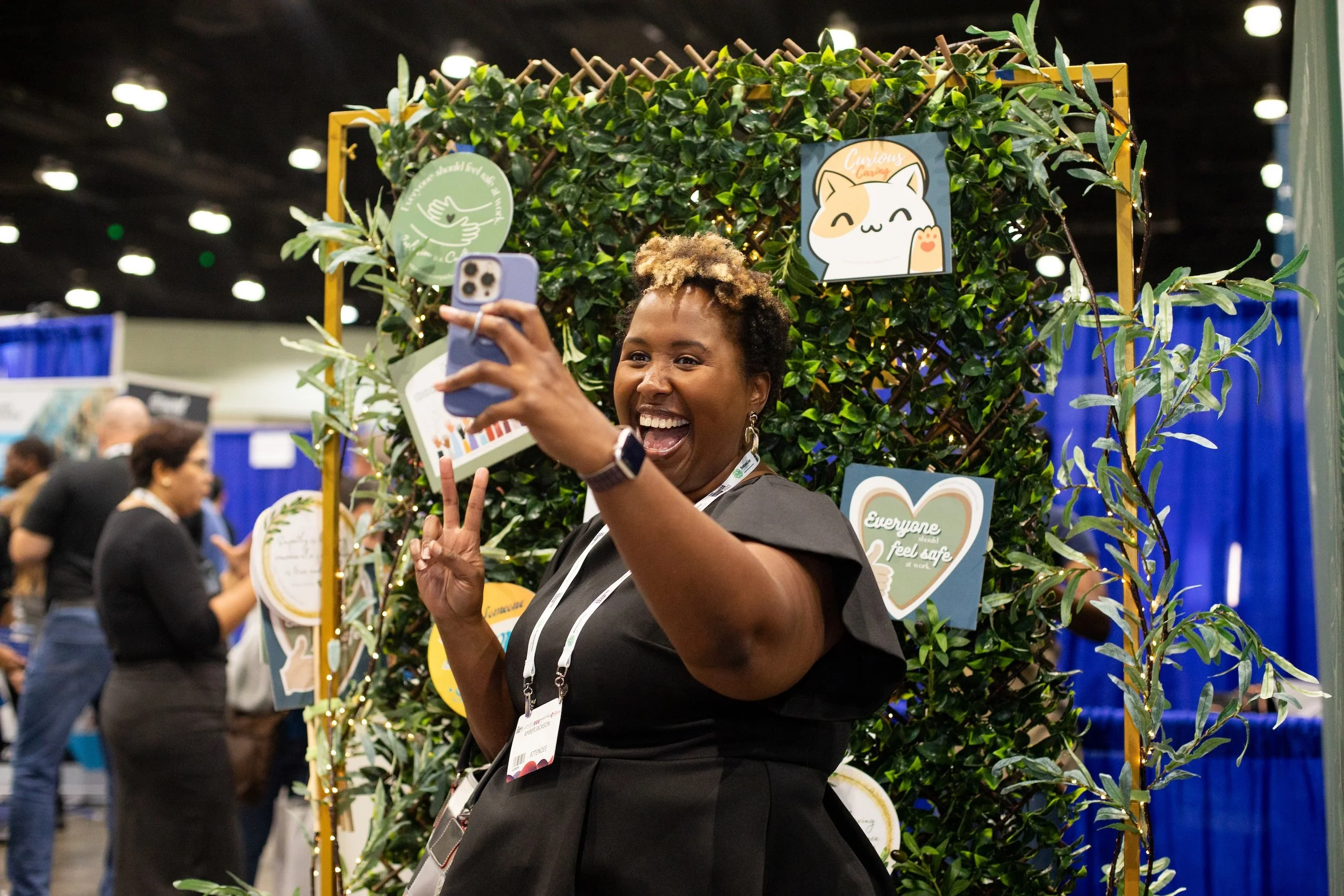 A woman taking a selfie at an event, standing in front of a decorative backdrop with green foliage and colorful signs. She is smiling, making a peace sign, and wearing a black dress.