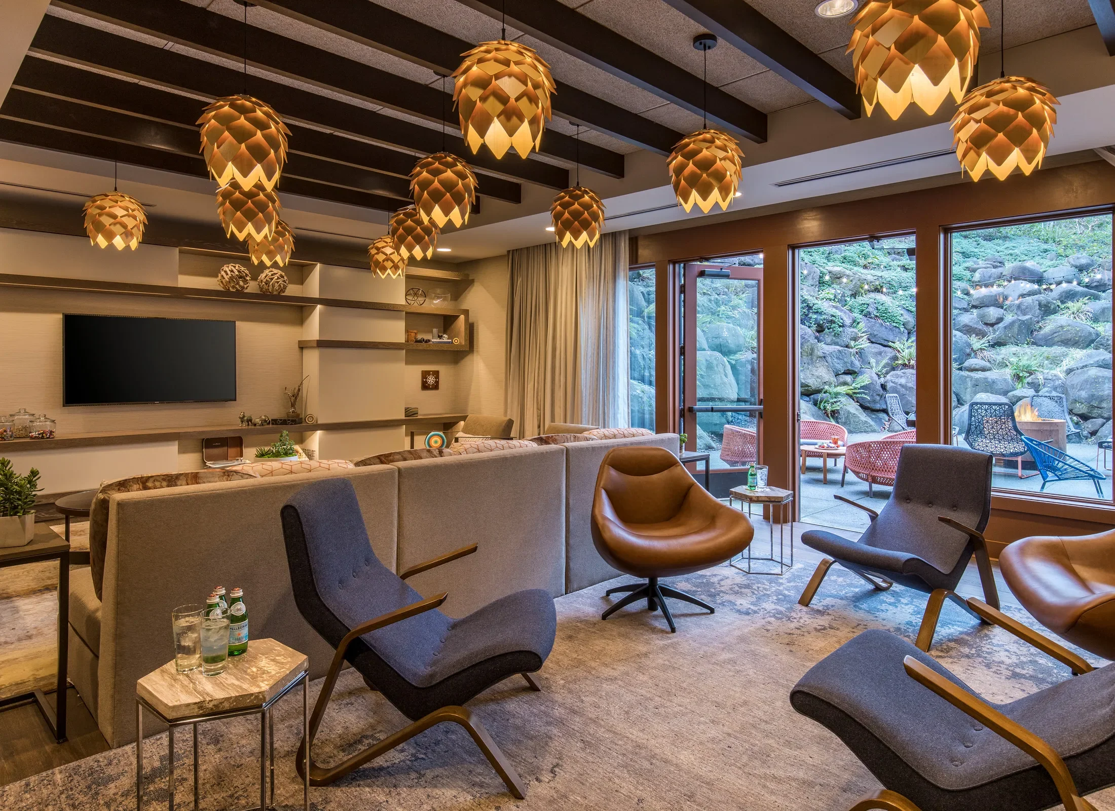Modern living room with mid-century style furniture, wooden ceiling beams, and large windows showing a rocky outdoor patio with seating, all illuminated by hanging pendant lights resembling pine cones.