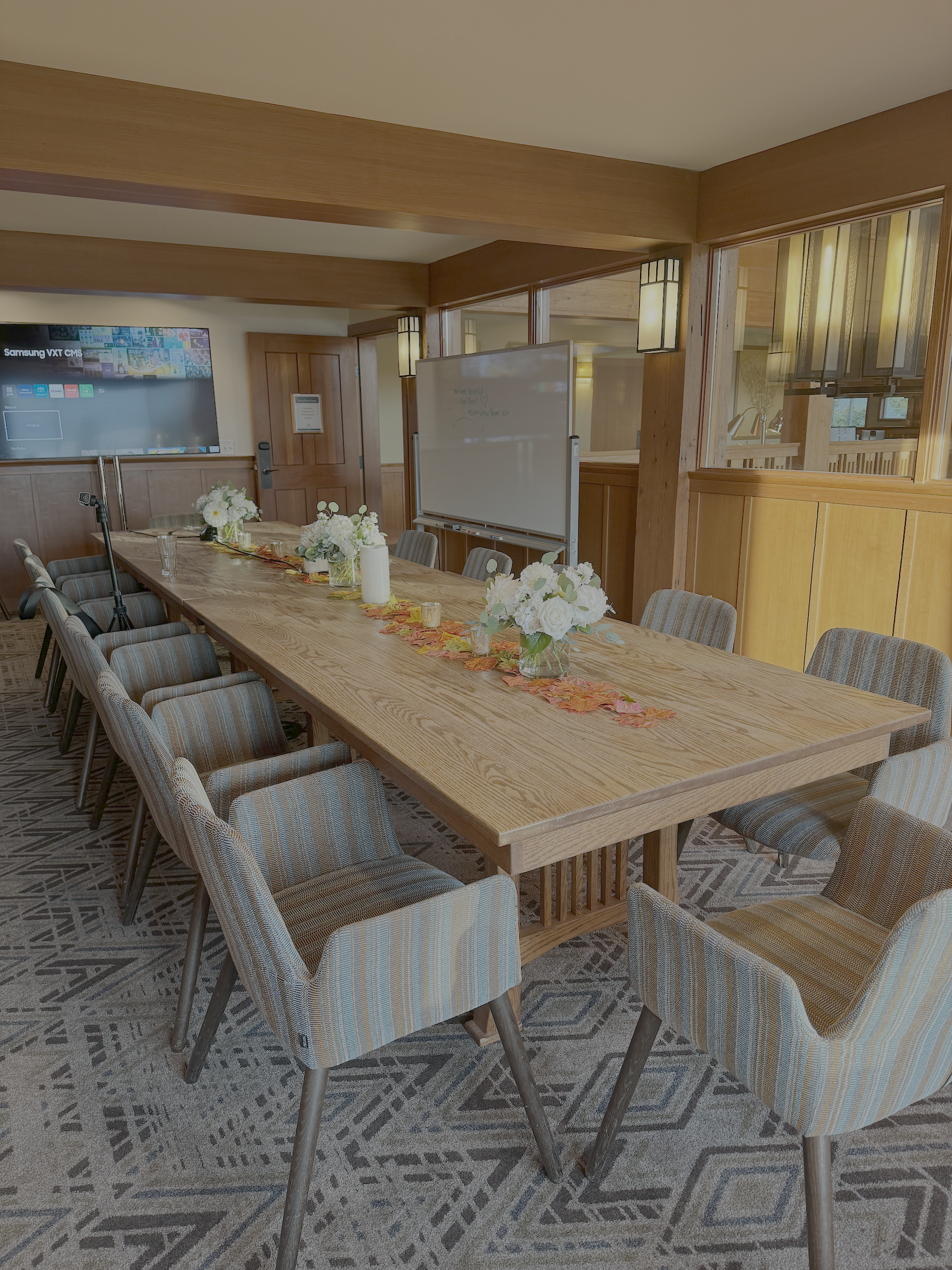 A conference room with a long wooden table adorned with white floral centerpieces, surrounded by upholstered chairs, and a whiteboard on a stand in front of wooden windows.