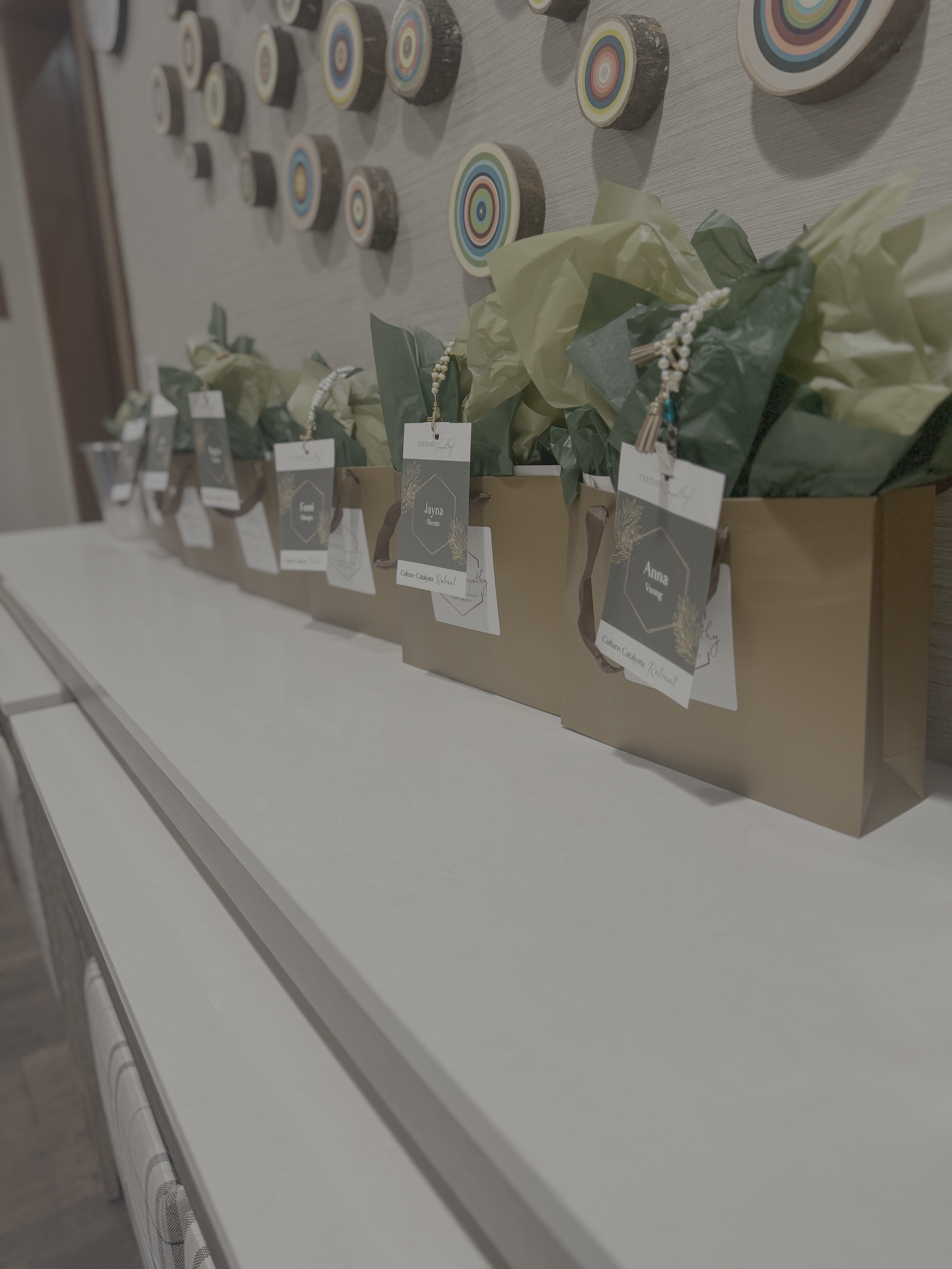 A display of wooden discs with colorful, concentric circle patterns, arranged on a wall above a table with gift bags wrapped in yellow and green tissue paper and tags with names.