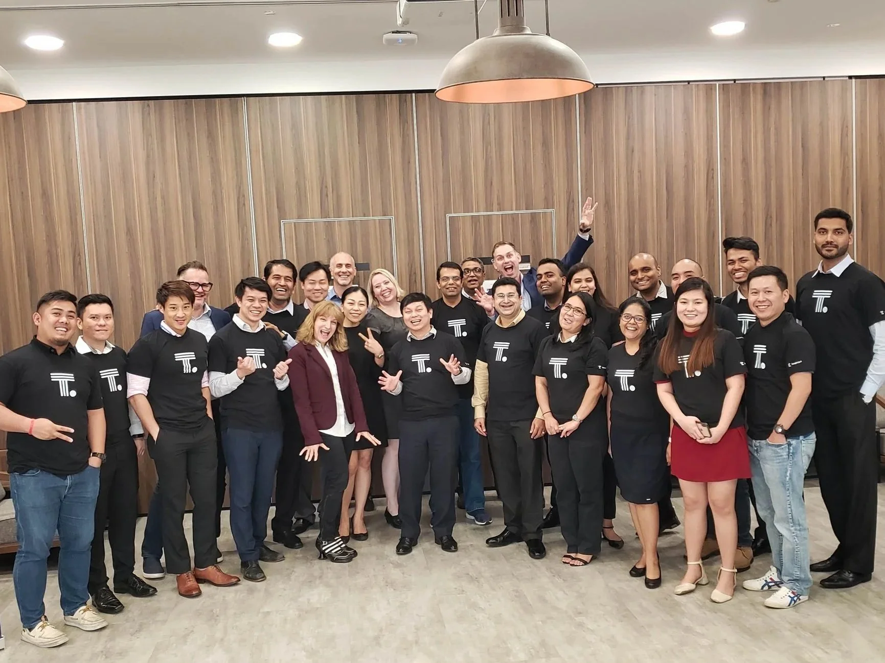 Group of diverse people posing for a photo indoors, some wearing matching black shirts with a white logo, smiling and displaying various gestures of happiness.