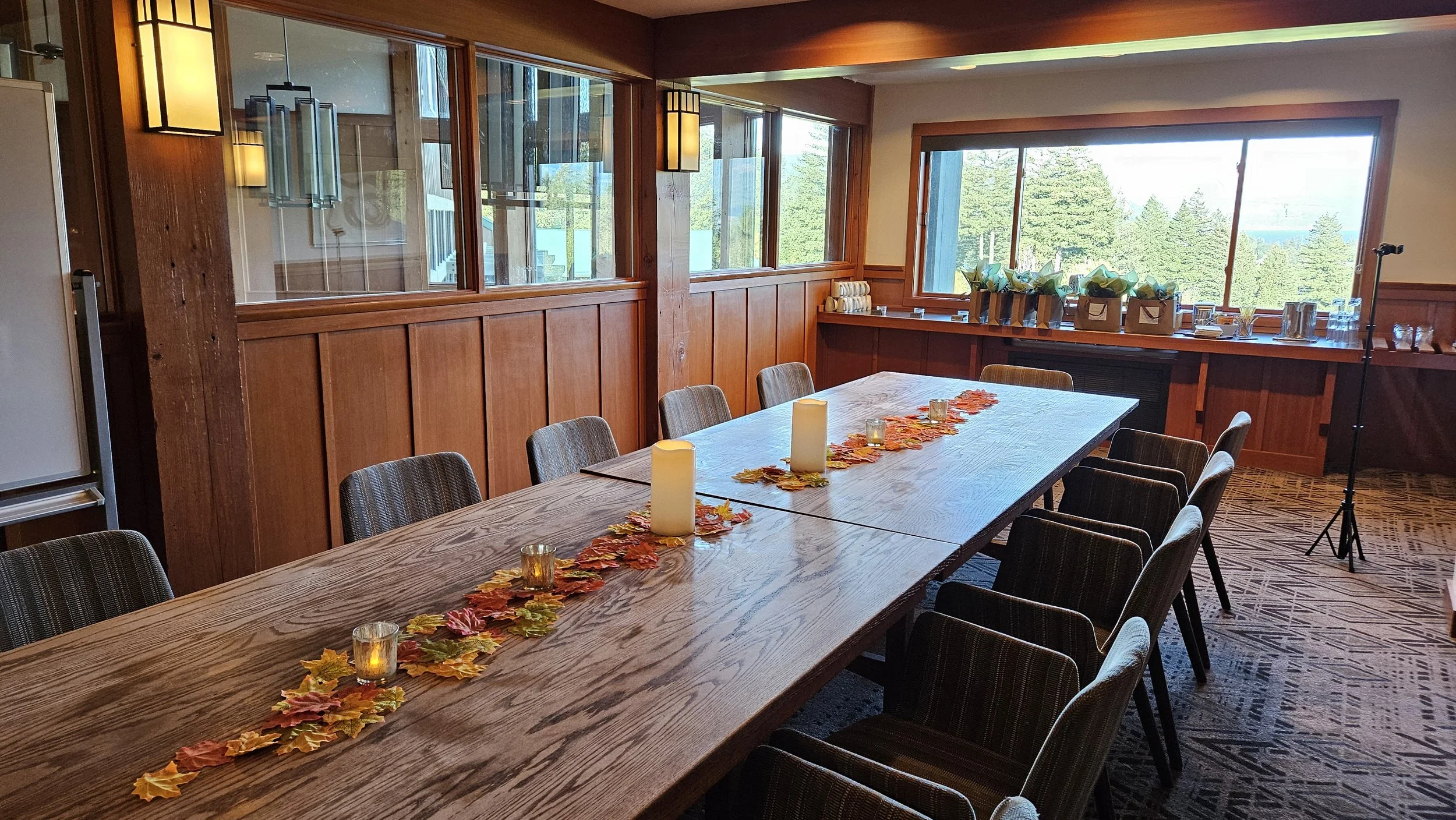 A long wooden dining table decorated with artificial autumn leaves, candles, and small votive holders, set in a room with wood-paneled walls and large windows showing a view of trees outside.