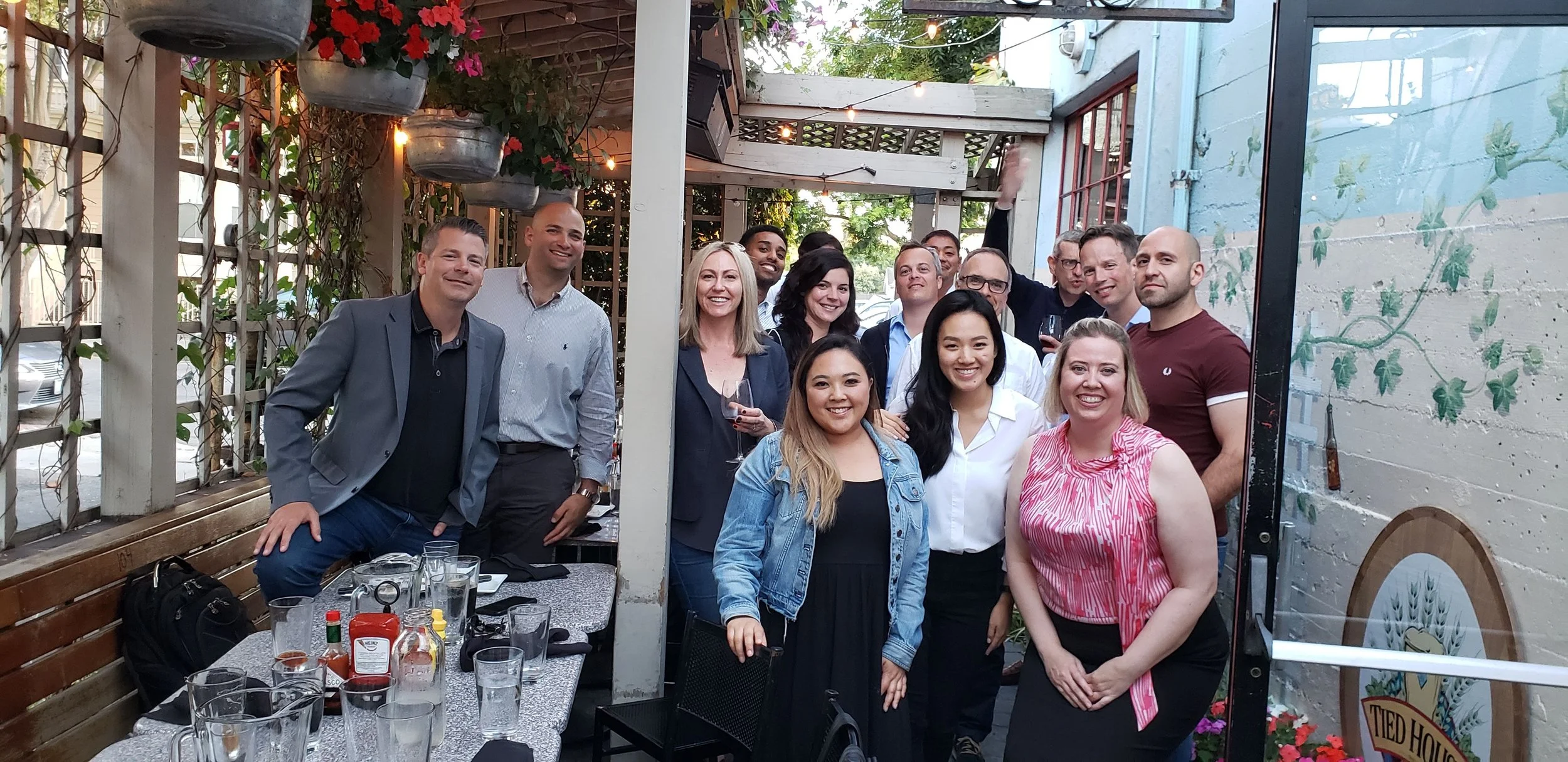 A group of people at an outdoor restaurant or patio, smiling and posing for a photo, with some holding drinks and condiments on the table.