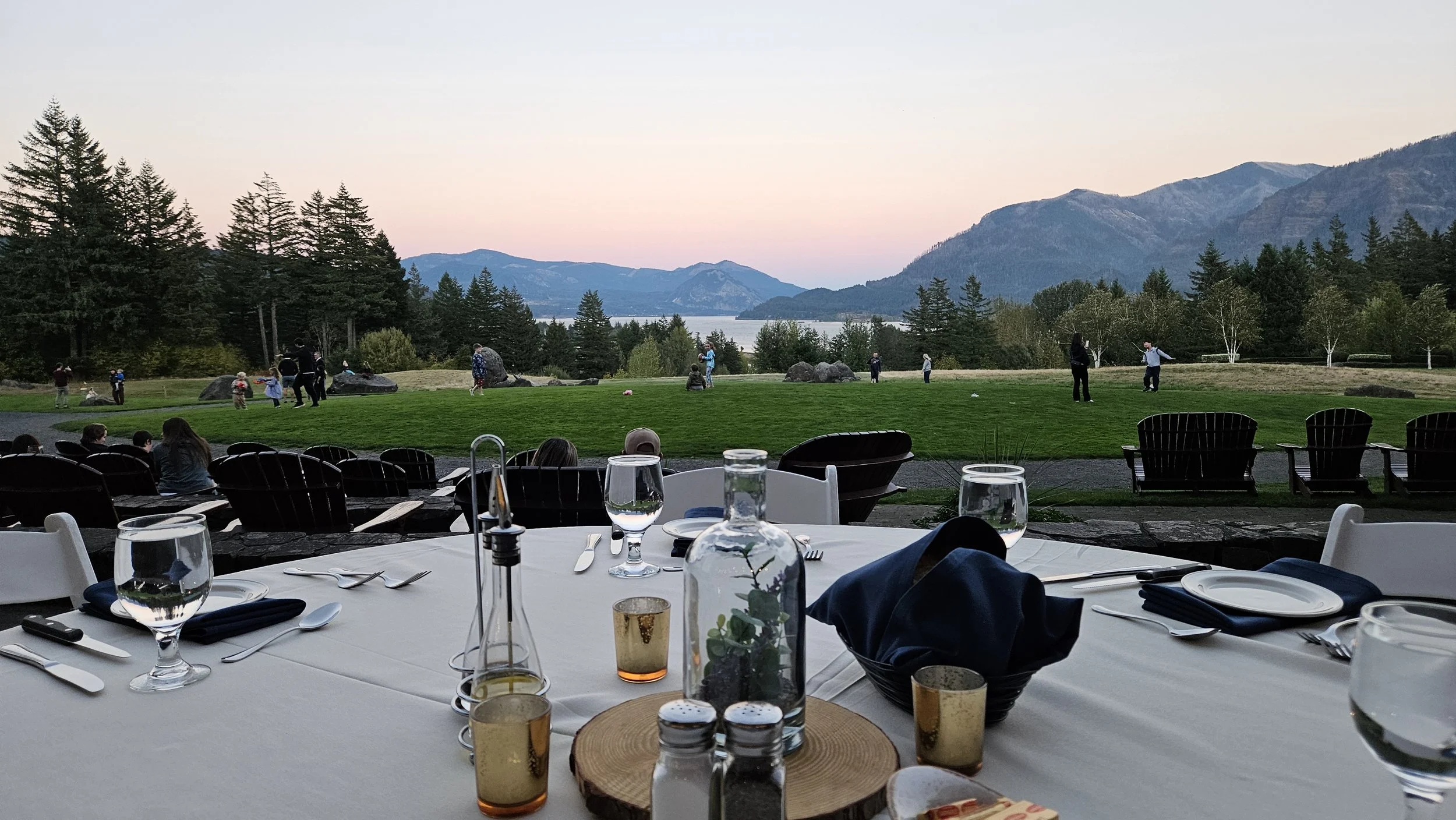 Dining table set with glasses, cutlery, and a bottle with a plant, overlooking a grassy outdoor area with people playing, surrounded by trees and mountains during sunset.