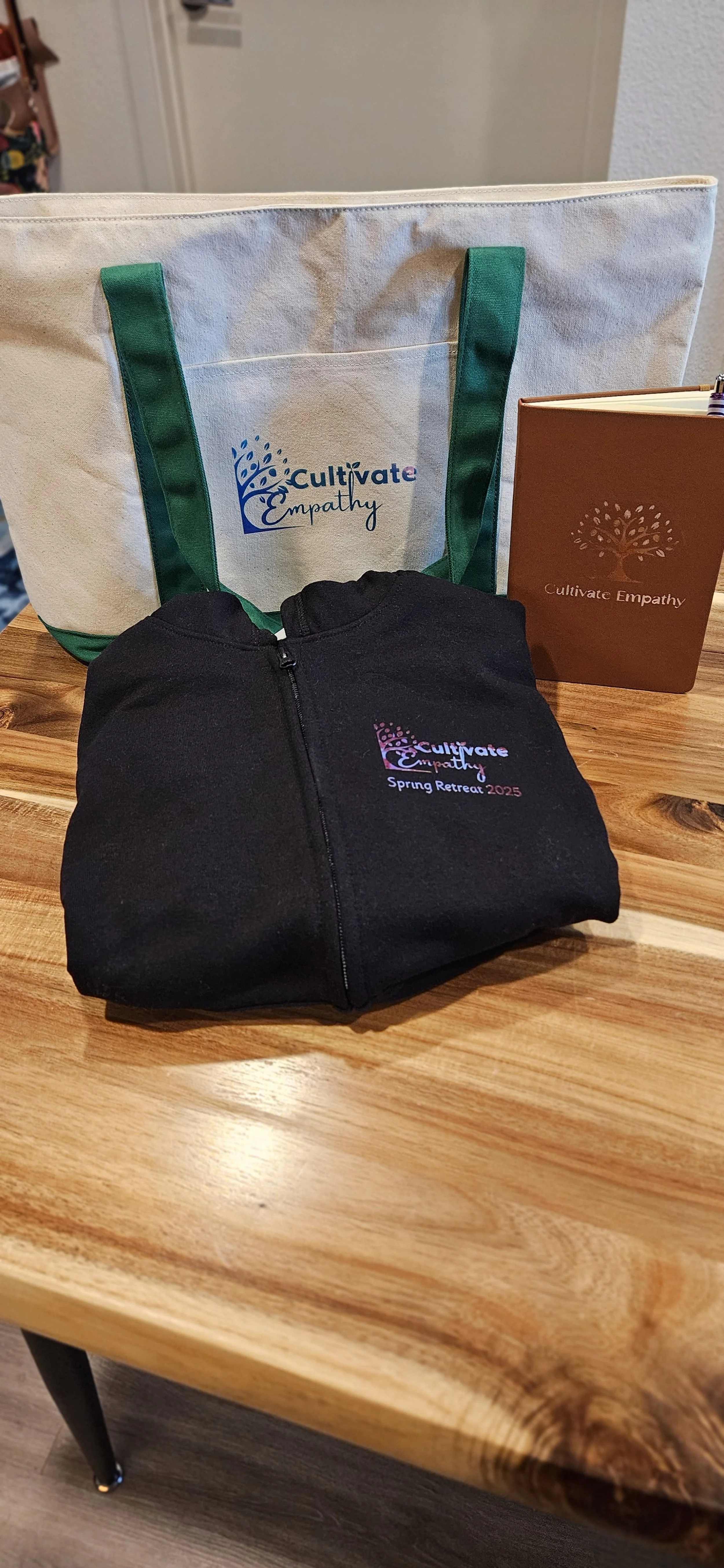 A wooden table displaying a white tote bag with green handles, a brown notebook, and a black zipped hoodie, all branded with 'Cultivate Empathy' and event details.