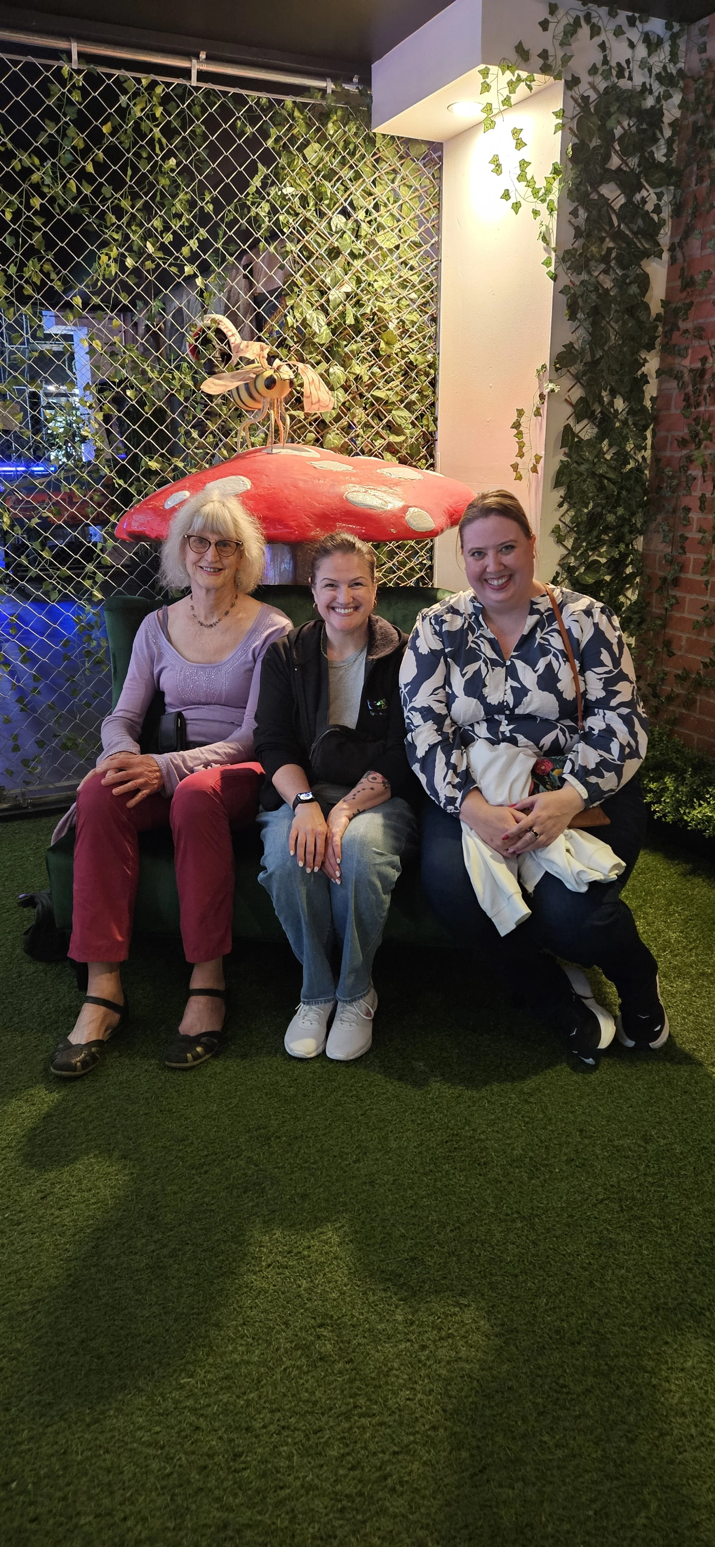 Three women sitting on a green sofa in front of a decorative setting with a large red and white mushroom, a bee decoration, greenery, and a brick wall. They are smiling and posing for the photo.