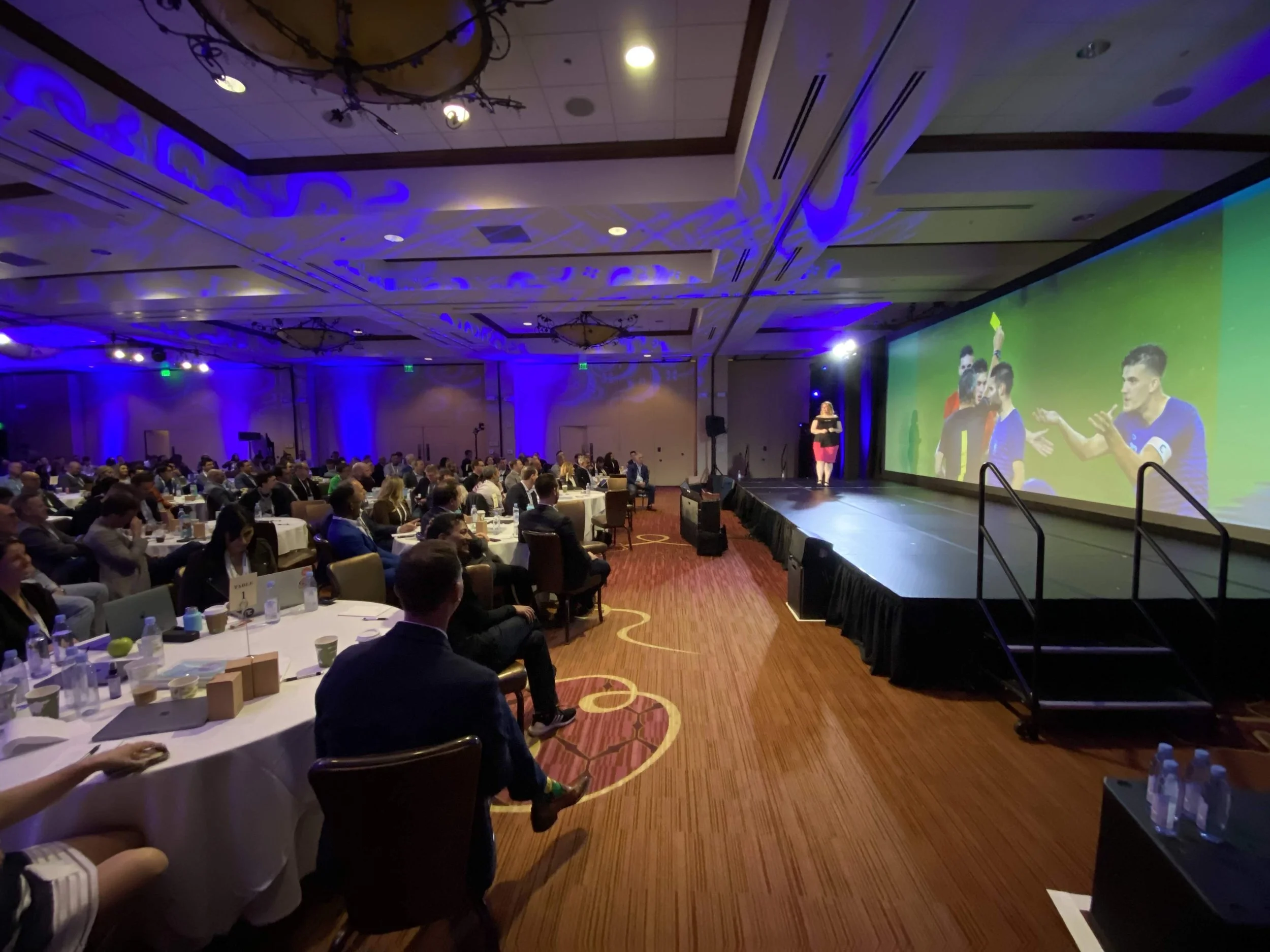 Large conference room filled with seated attendees facing a stage and a large screen. The stage has a speaker and a woman presenter. The room is decorated with blue lighting and chandeliers, and has a red patterned carpet.