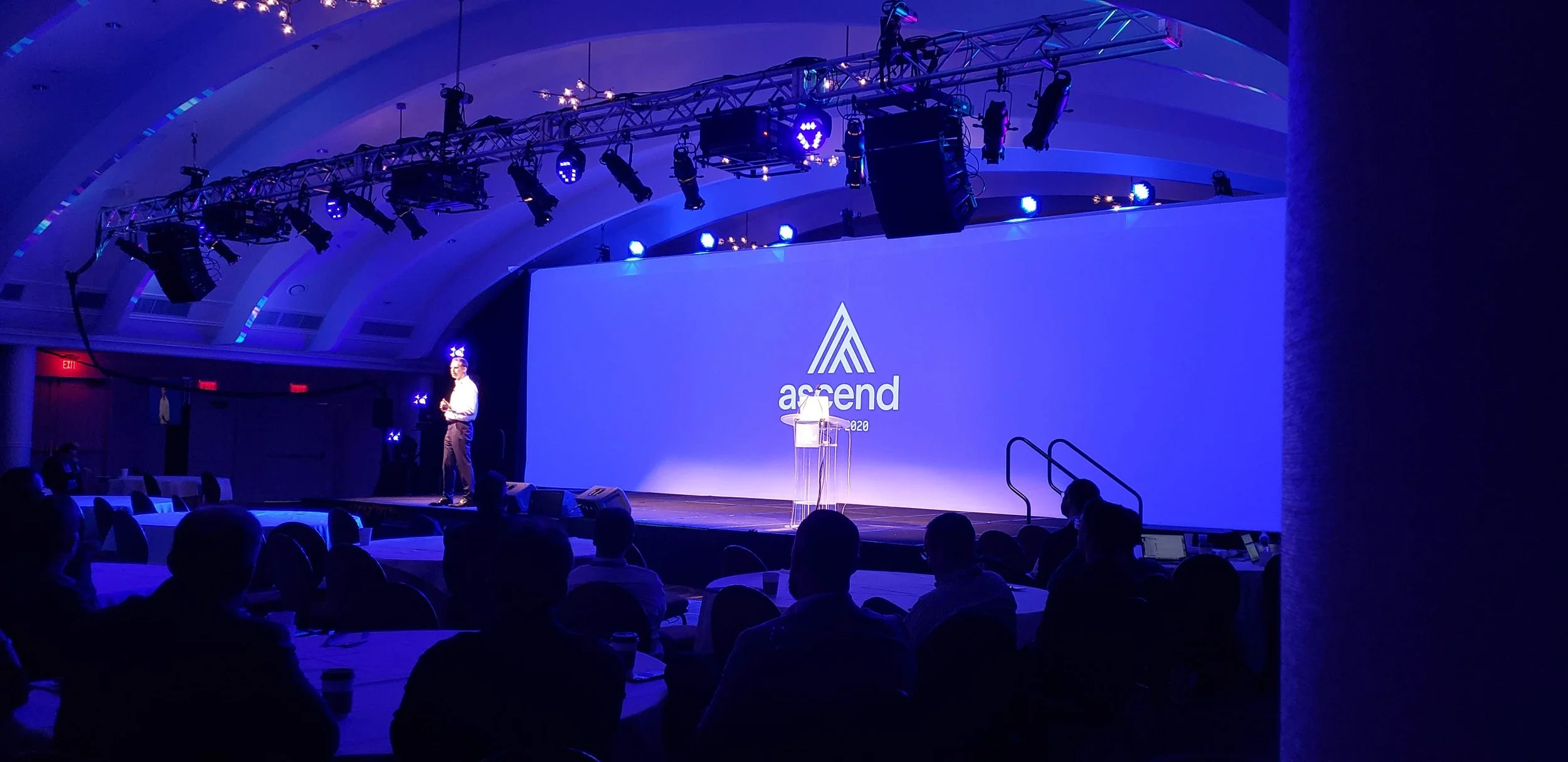 A large conference room with a stage, projection screen displaying the 'ascend' logo, and a speaker presenting at a podium under blue and purple lighting.
