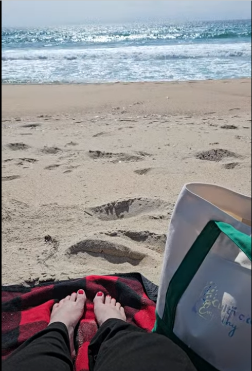 Person relaxing on a beach towel in the sand, with footprints nearby, facing the ocean with waves and a cloudy sky.