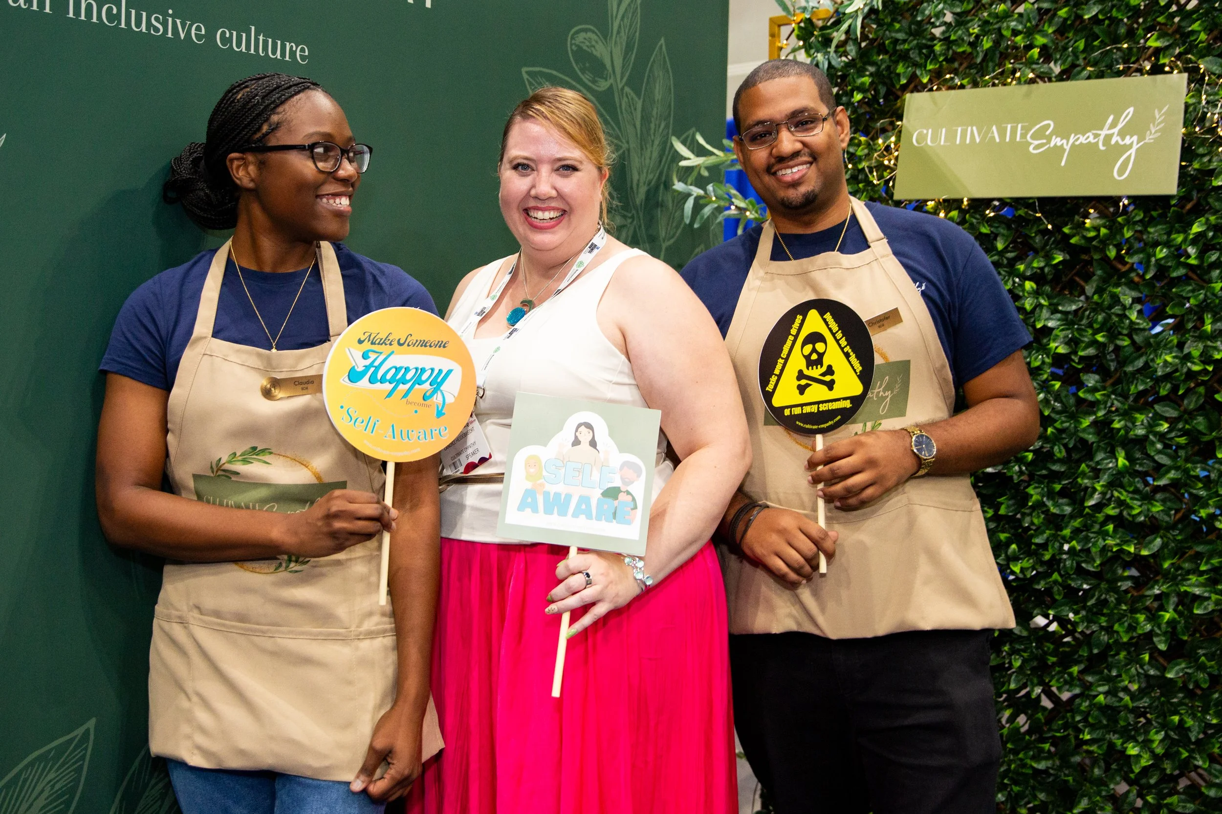 Three diverse people standing together at an event, smiling and holding signs that promote self-awareness and empathy, with a green themed background that includes the words 'Cultivate Culture' and 'Cultivate Empathy'.