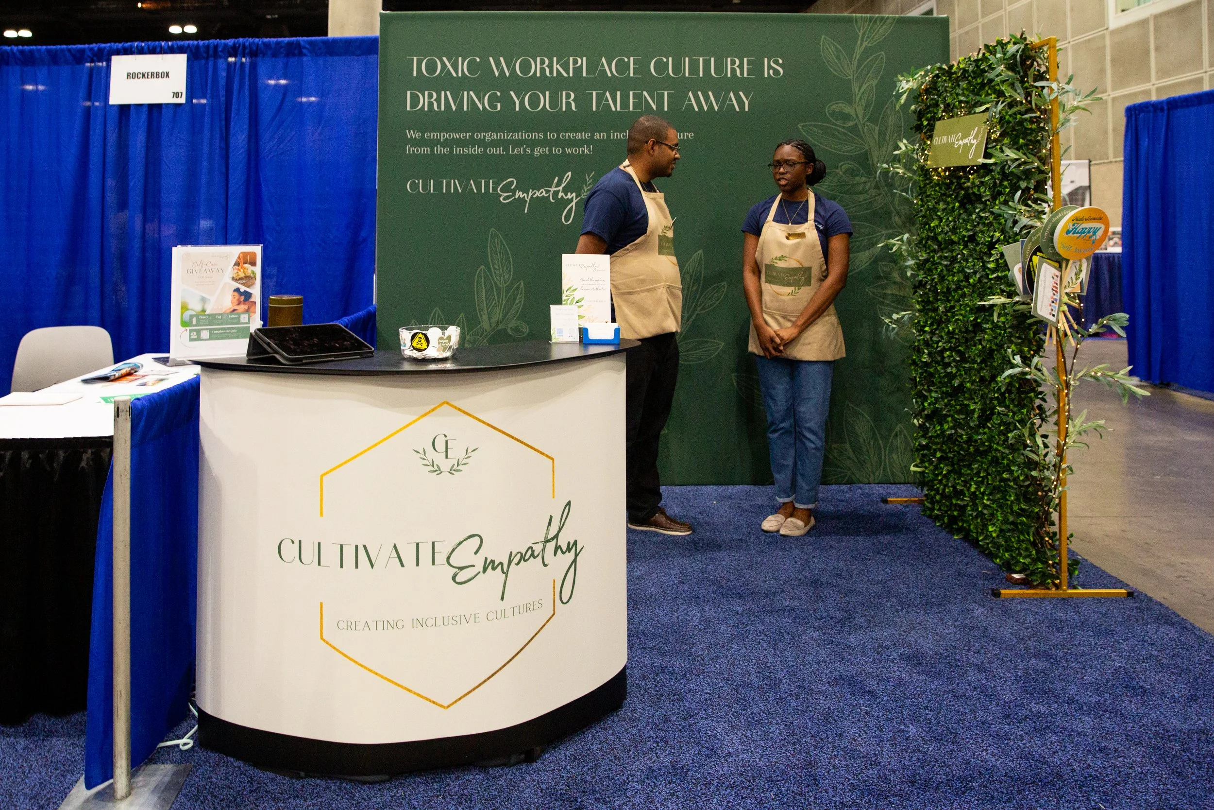 Booth at a conference for Cultivate Empathy, with a green backdrop and two people talking. The booth features a white curved counter with the Cultivate Empathy logo, a small table to the left with promotional materials, and a decorative green plant o