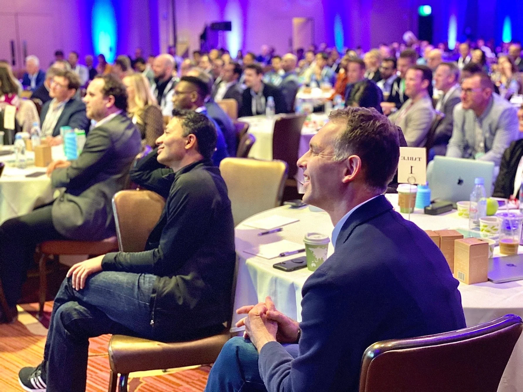 Large conference room filled with a diverse group of people seated at round tables, listening and smiling, with some taking notes. The room is illuminated with purple and blue lighting.