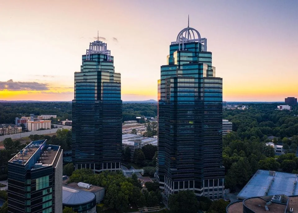 Aerial view of two modern skyscrapers with dark glass facades and unique architectural tops in Sandy Springs, Georgia, set against a sunset sky in an urban area with trees and surrounding buildings.