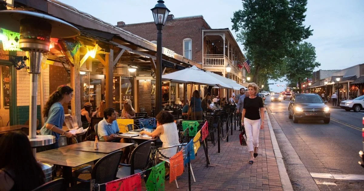 People dining outside at a restaurant on a lively city street in Roswell, Georgia, during dusk. Spacious sidewalk with outdoor tables, some under umbrellas, and a woman walking along the sidewalk. Buildings with balconies and string lights.