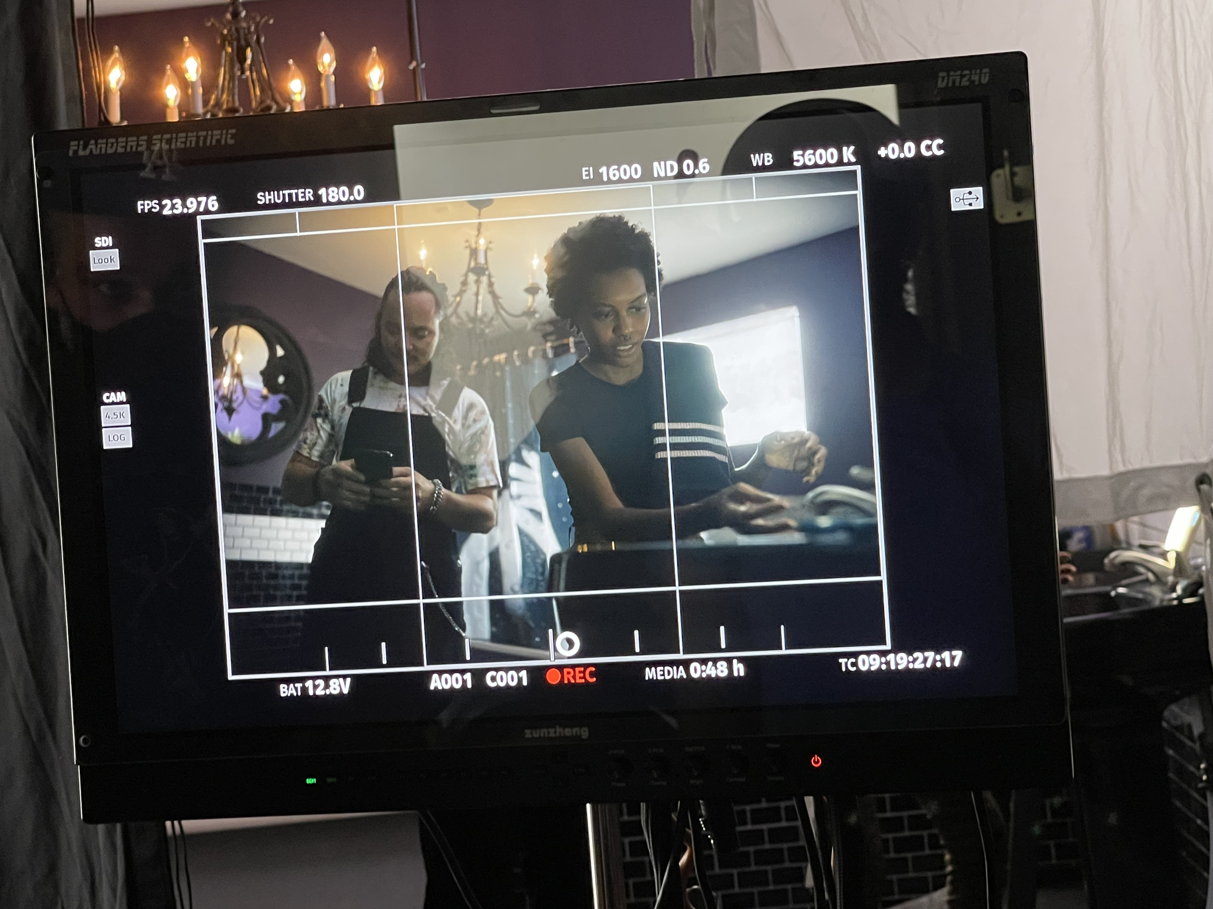 Two women on a film set, one is looking at her phone and the other is handling a script or a document, viewed through a camera monitor.
