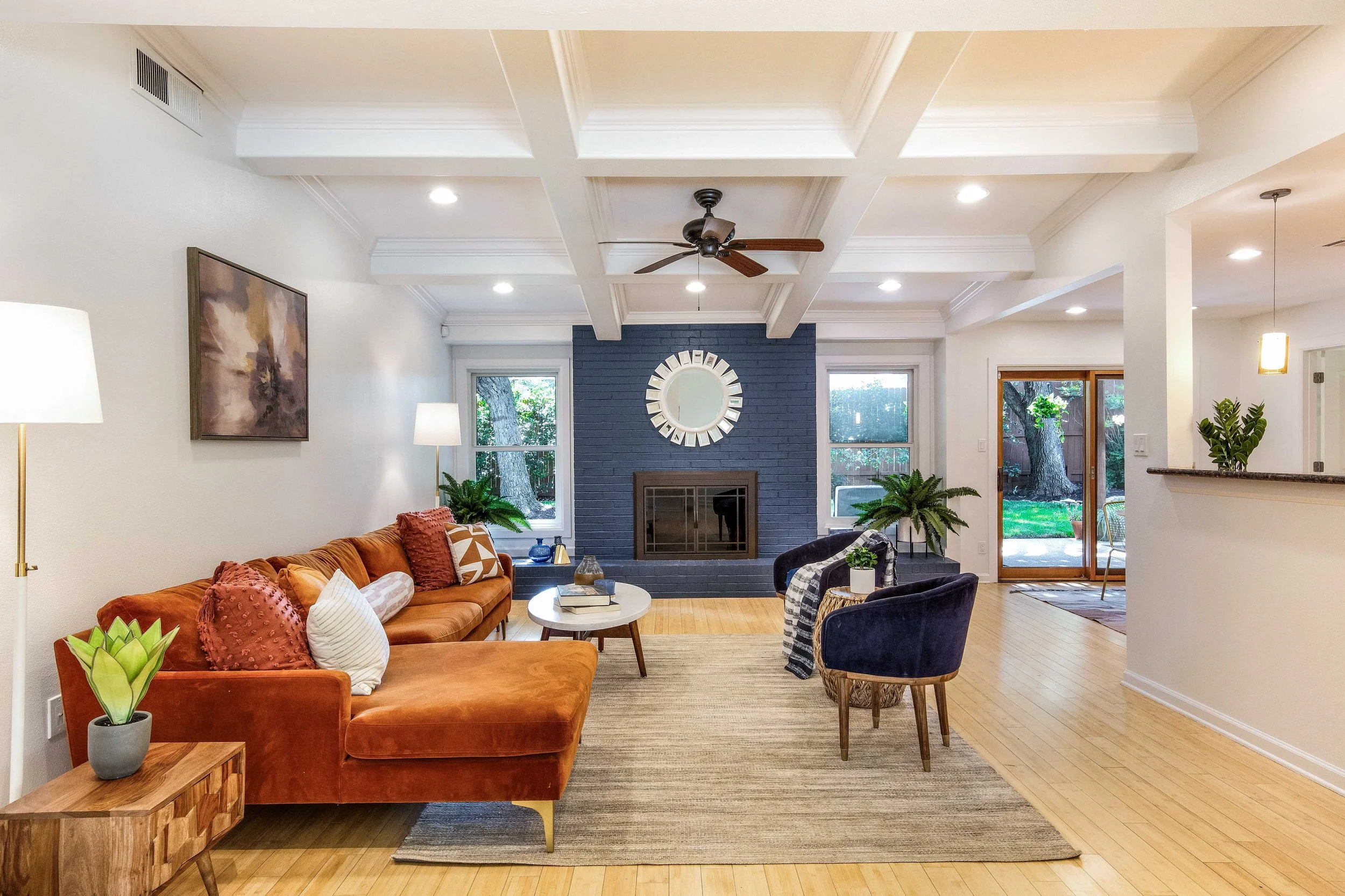 Living room with white walls, coffered ceiling, and hardwood floors. Fireplace with a dark blue brick wall, round mirror above it, and large windows on either side. Two dark blue armchairs and a rust-colored sofa with pillows. Small coffee table and 