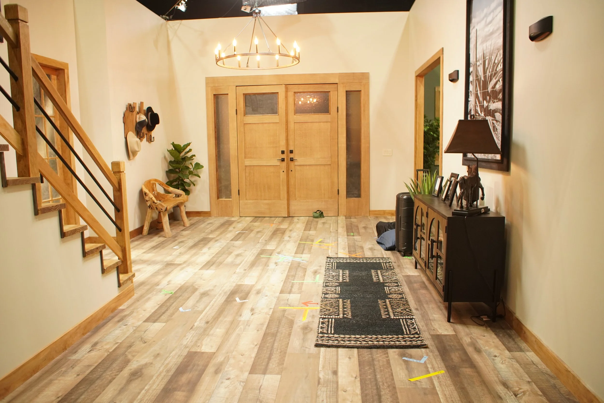 Empty living room with wooden flooring, a black and cream area rug, a black sideboard with family photos, a table lamp, and a green plant. Wooden stairs lead to the upper level, and decorative hats hang on the wall next to a wooden bench. Double wood