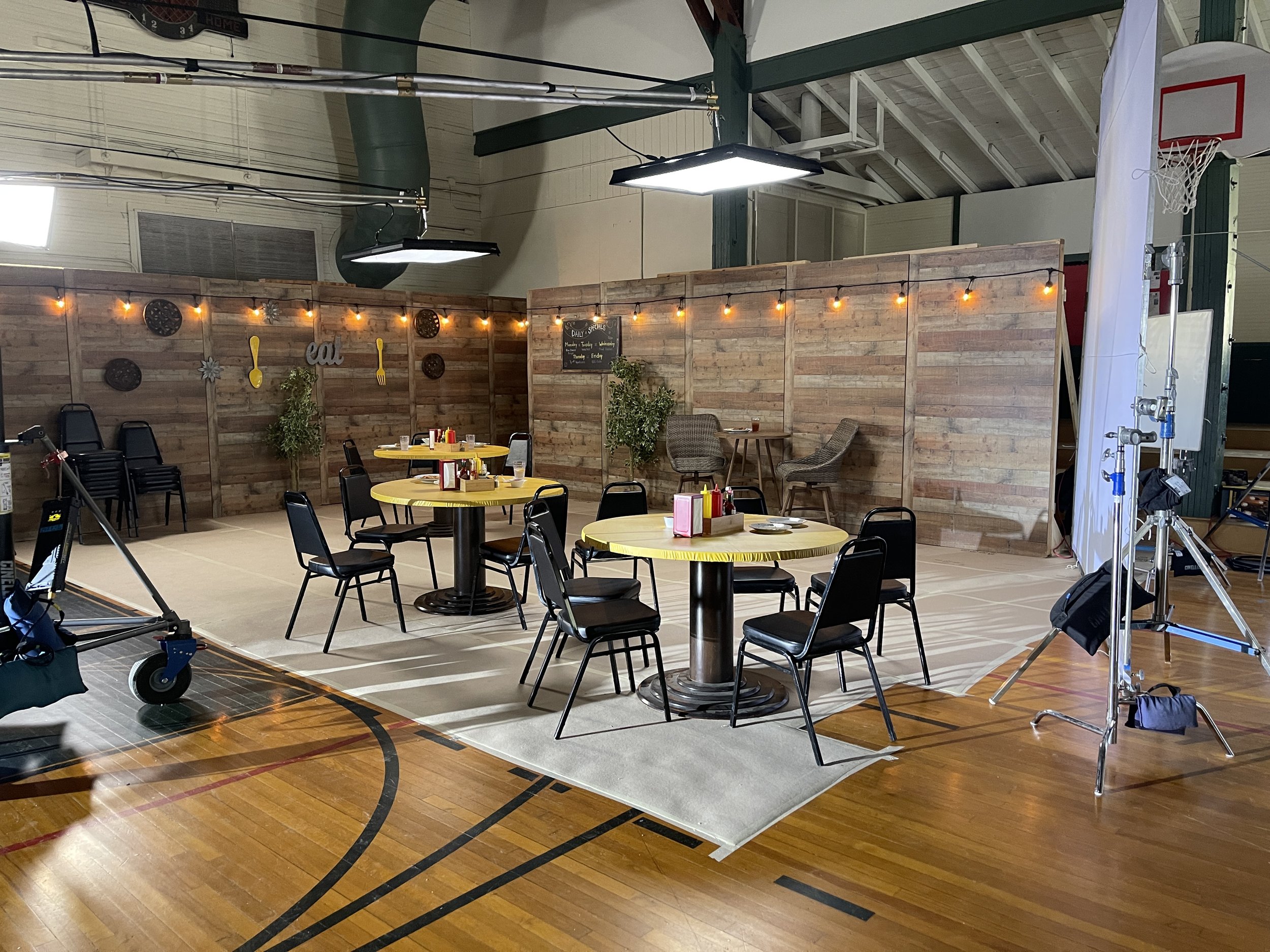 Empty indoor event space set up for a casual gathering, with round yellow tables, black chairs, a wooden backdrop decorated with string lights and wall art, a small chalkboard menu, and stage lighting equipment.
