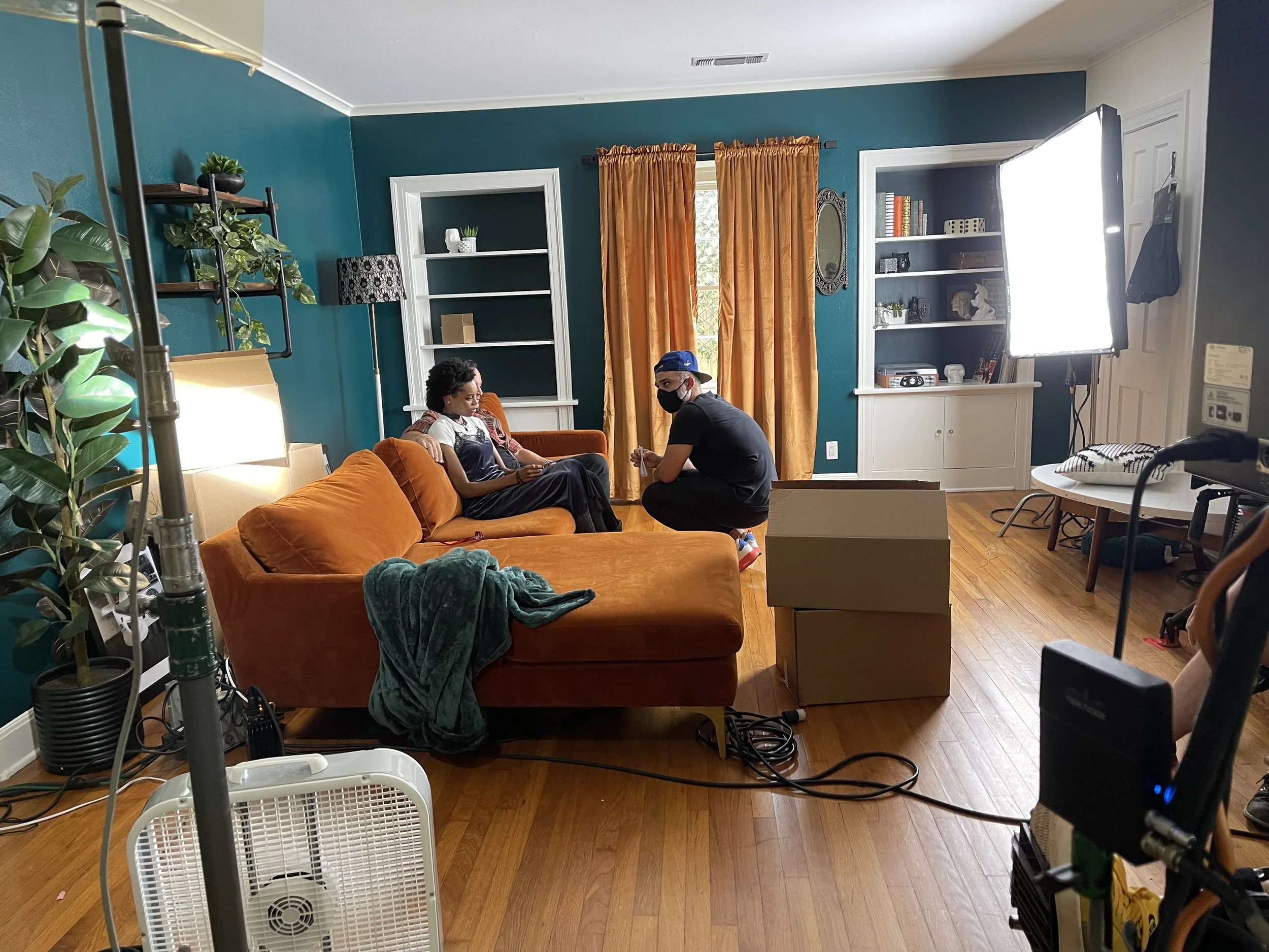 A person with curly hair sitting on an orange sofa, another person with a mask crouching in front of them, and a crew member with a blue cap and dark clothing adjusting equipment, in a living room set with a large light on right side, a fan and camer