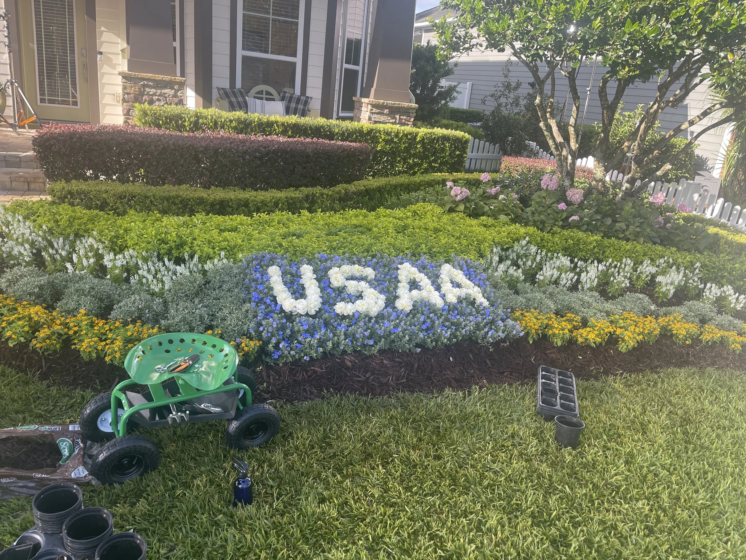 A well-maintained garden with colorful flowers spelling out 'USA' and various plants, shrubs, and a small tree, with gardening tools and supplies in the foreground.