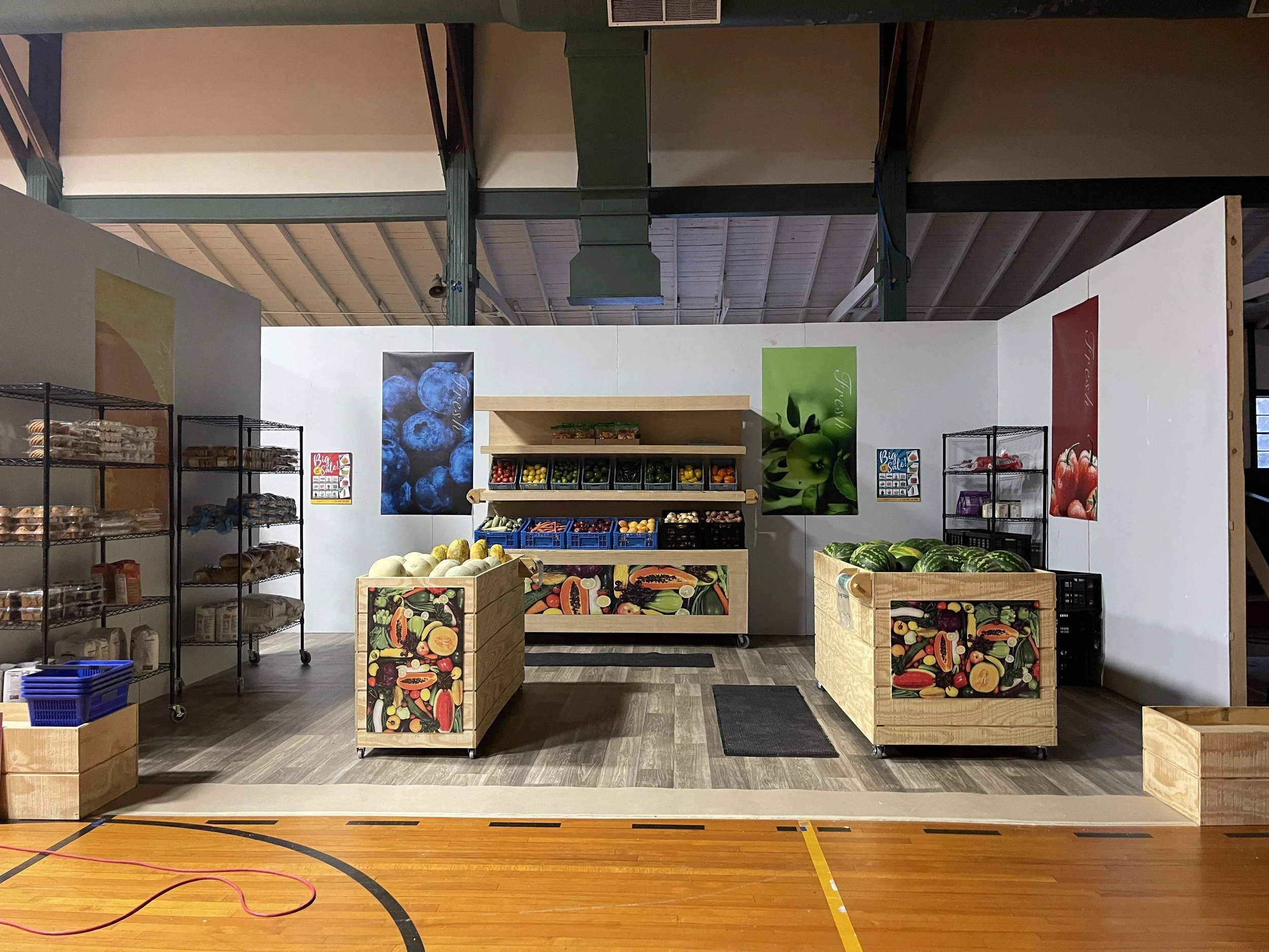 Small grocery store display with wooden crates and metal shelves holding fresh produce like watermelons, apples, melons, and other fruits and vegetables, set against a white wall with colorful fruit posters.