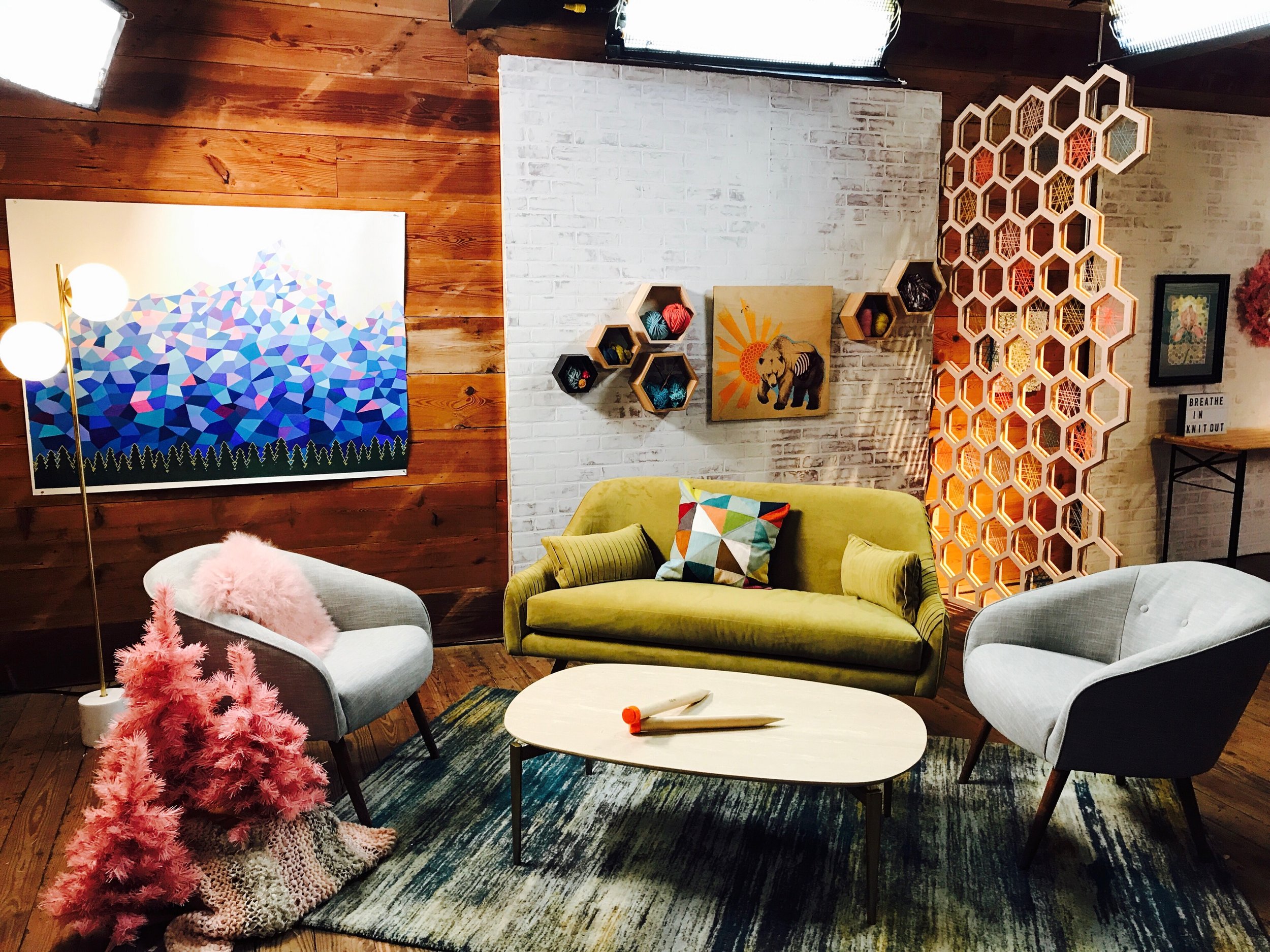 Cozy living room with mid-century modern furniture, including a green sofa with colorful cushions and two light gray armchairs. There is a small pink Christmas tree, a white oval coffee table with a white and orange object, and a geometric hexagon-pa