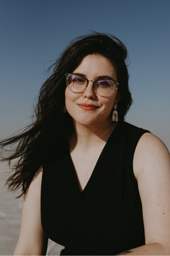 Woman with dark hair and glasses smiling at the camera outdoors against a clear blue sky.