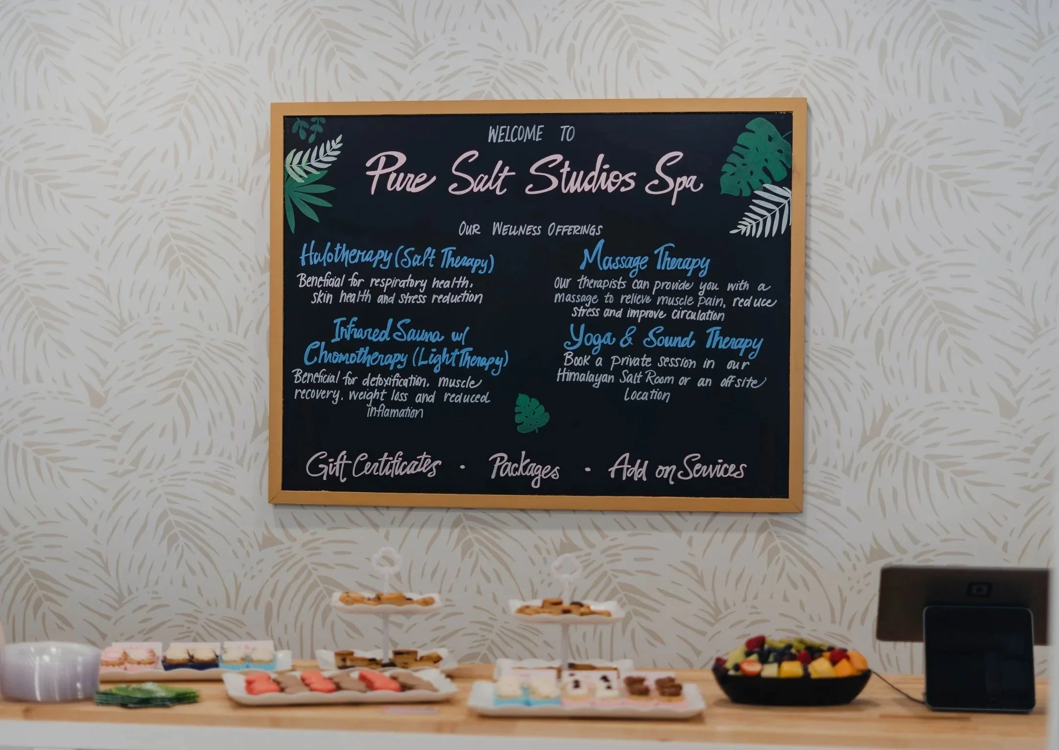 A blackboard hanging on a wall with a list of wellness offerings at Pure Salt Studios Spa, including salt therapy, massage therapy, infrared sauna with chromotherapy, and yoga and sound therapy. Events at Pure Salt Studios