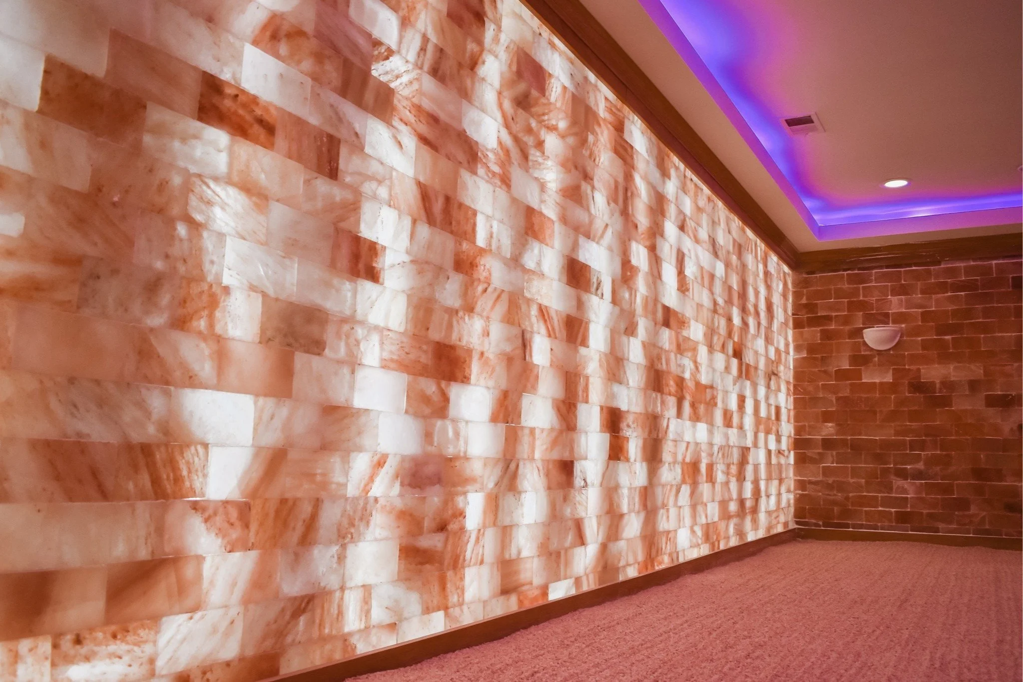 Interior of a room with a wall made of pink Himalayan salt bricks, brick wall, carpeted floor, and purple accent lighting in the ceiling.