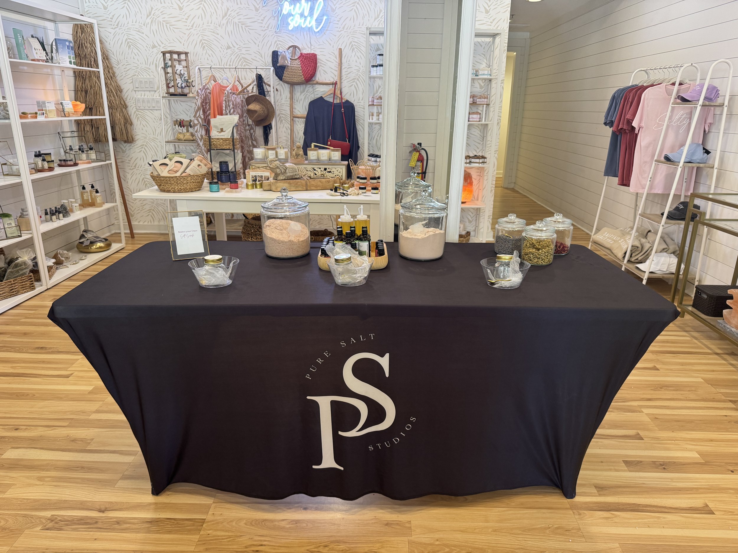 A retail display table covered with a black tablecloth, branded with 'Pure Salt Studios,' showcasing jars of salts and oils, with a store background featuring clothing and accessories on shelves and racks.