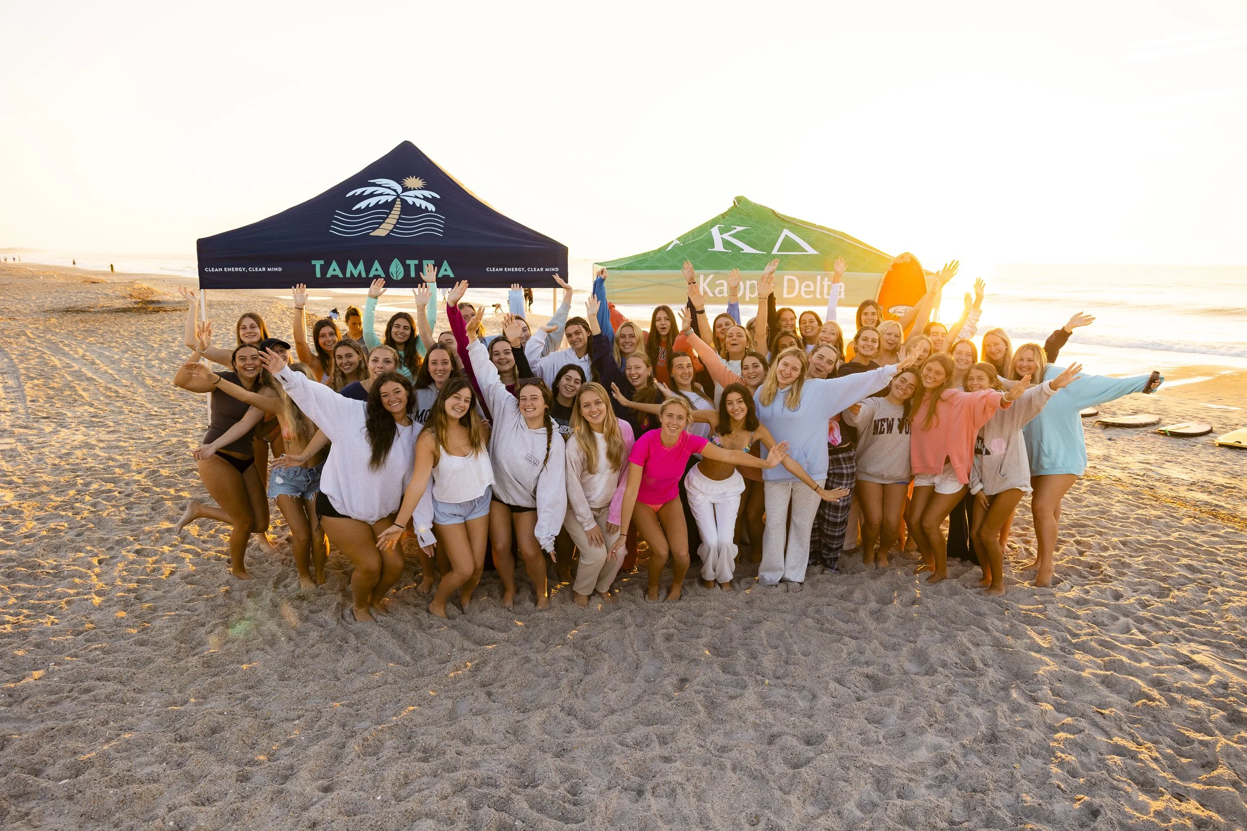 Group of young women in beachwear on the beach at sunset, smiling, waving, and posing in front of tents with Greek letters and logos.