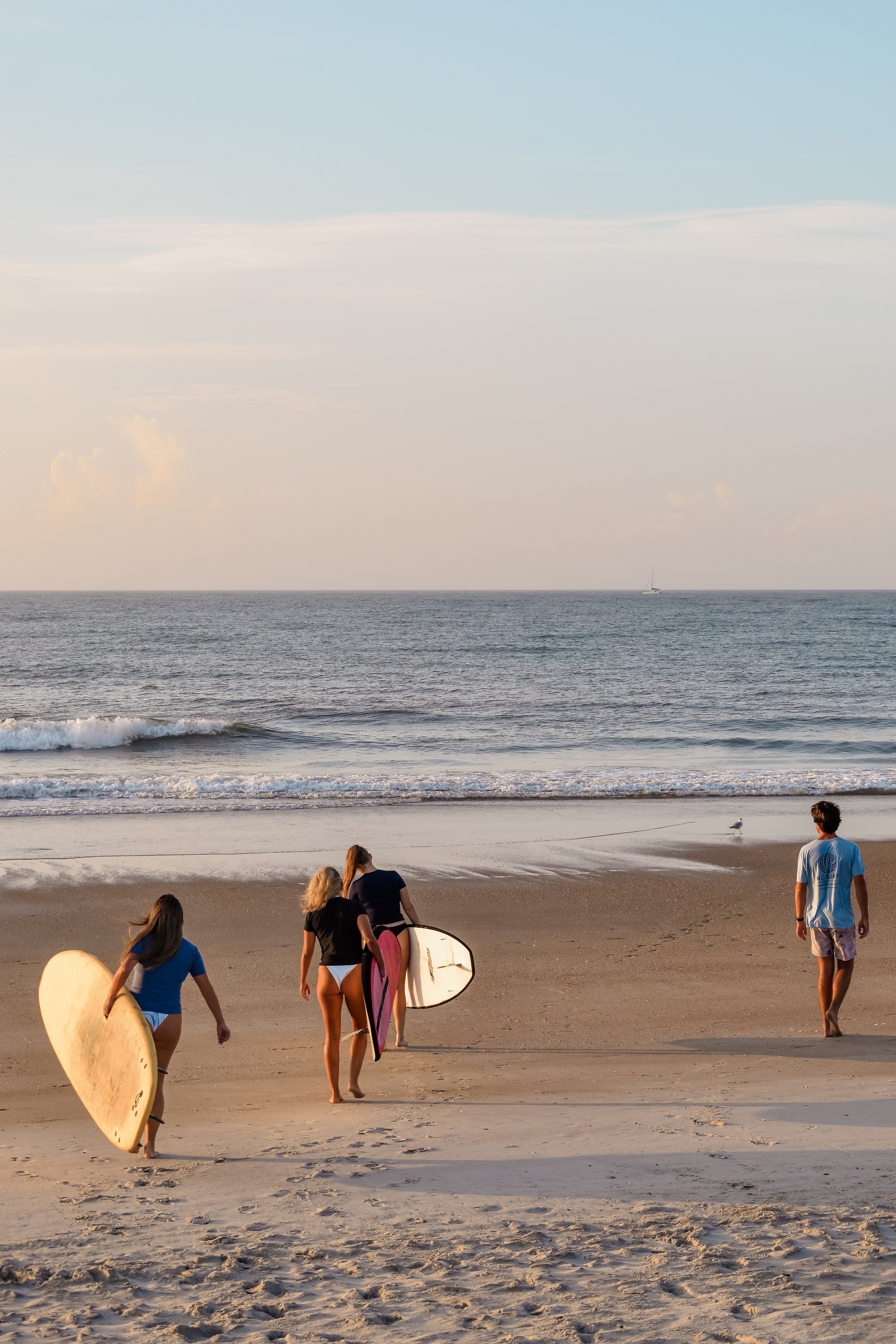 Four people, two women and two men, walking on a sandy beach with surfboards, heading toward the ocean at sunset.