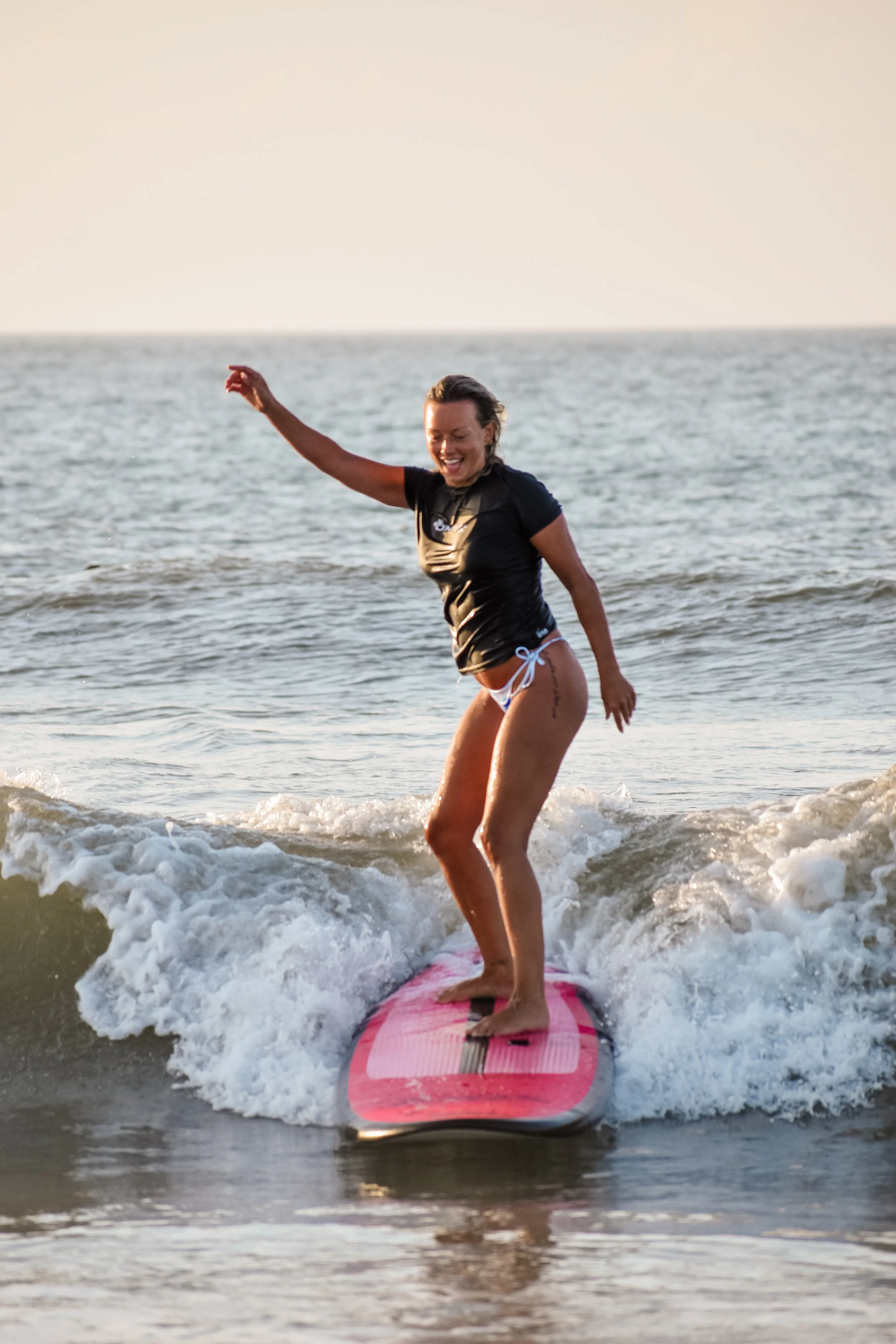 A woman smiling while surfing on a pink surfboard in the ocean with small waves, wearing a black T-shirt and white bikini bottoms.