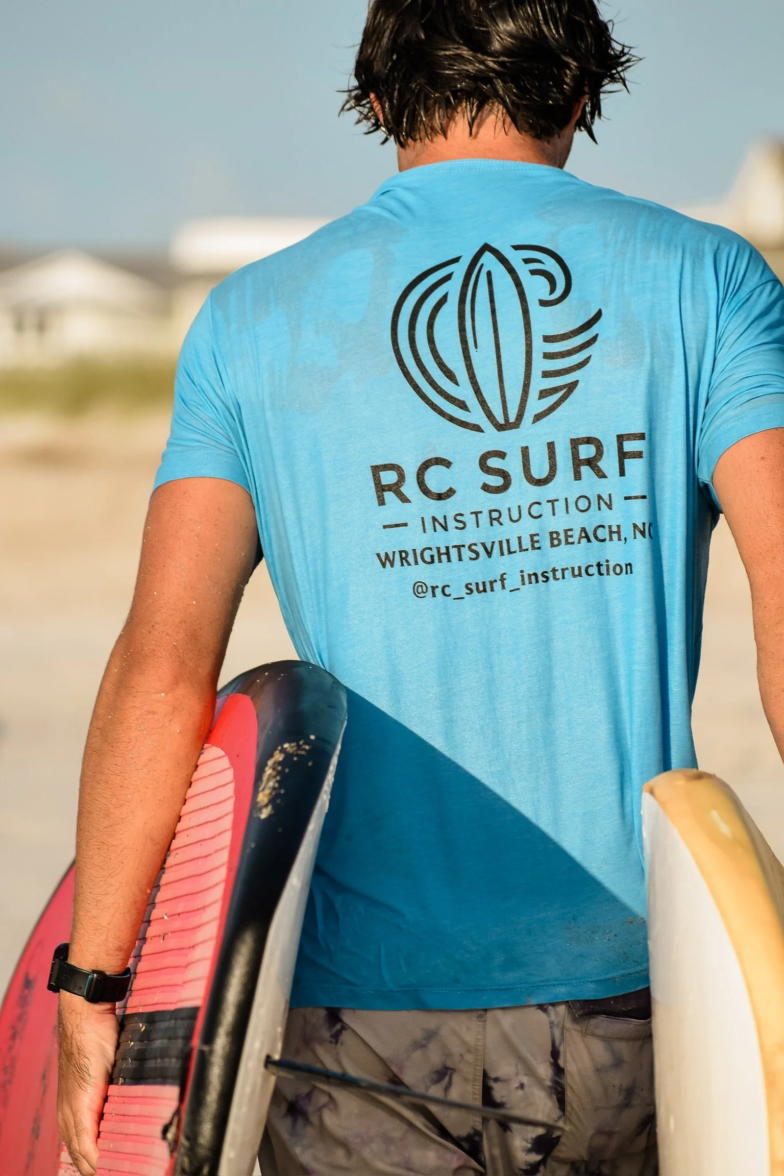 A person holding a surfboard at the beach, wearing a blue RC Surf instruction t-shirt, with houses and the beach in the background.
