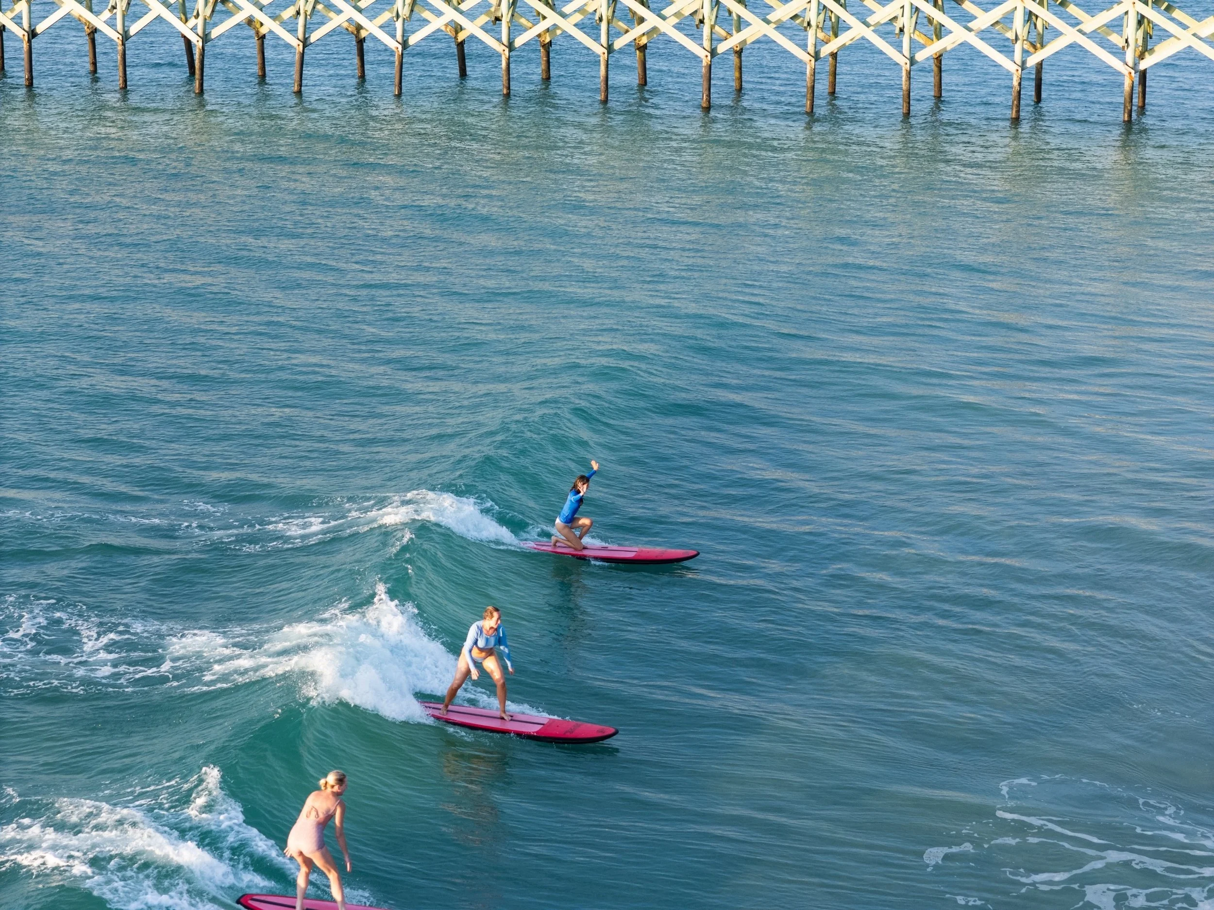 Three people riding red surfboards on the ocean near a pier.