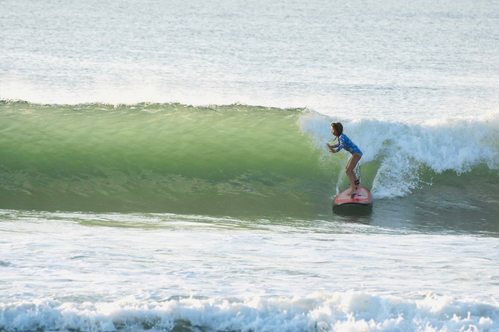 A young girl surfing on a pink surfboard in the ocean, riding a green wave.