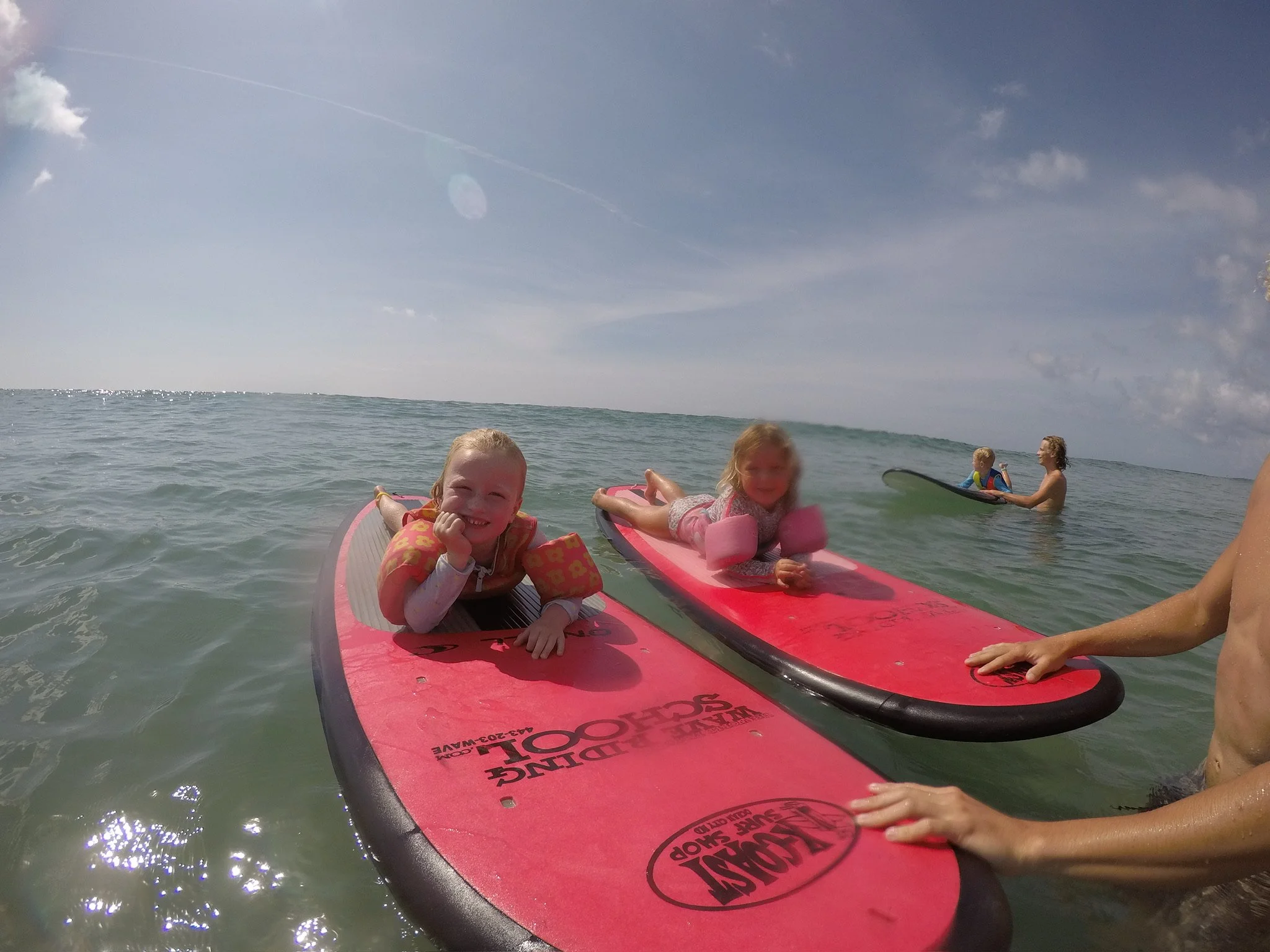 Two young girls are lying on pink paddleboards floating on the water, smiling at the camera, with a woman in the background holding a small child on another paddleboard and a man holding a paddle nearby.