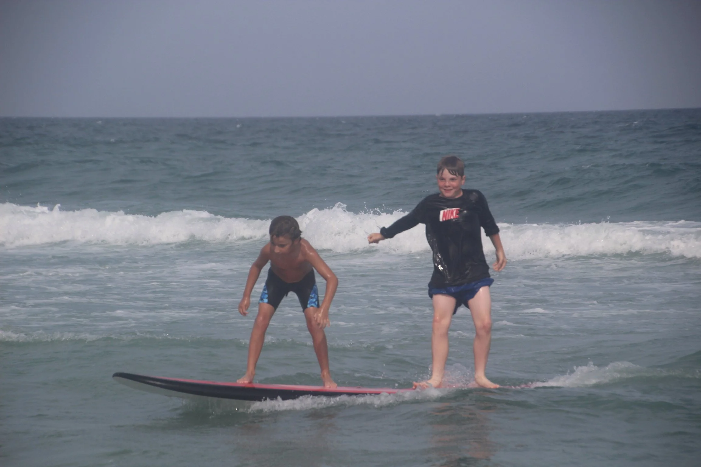 Two boys surfing on a surfboard in the ocean with waves and gray sky.