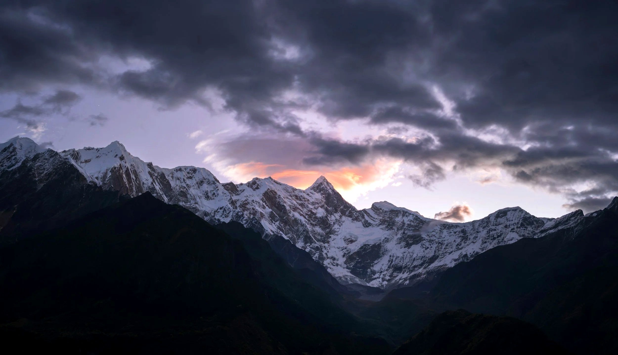 Snow-capped mountain range under a dark, cloudy sky with a hint of sunset light