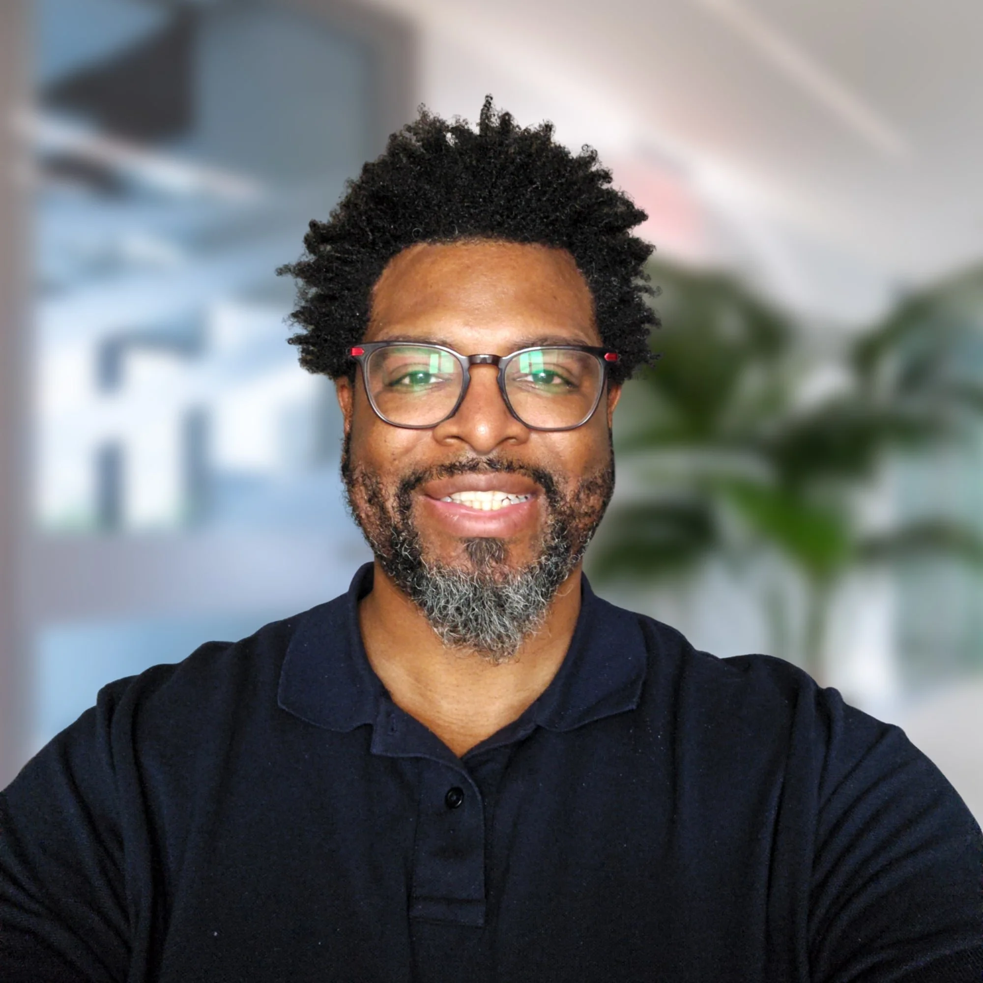 Close-up of a smiling African American man with glasses, black curly hair, and a beard, wearing a dark collared shirt.
