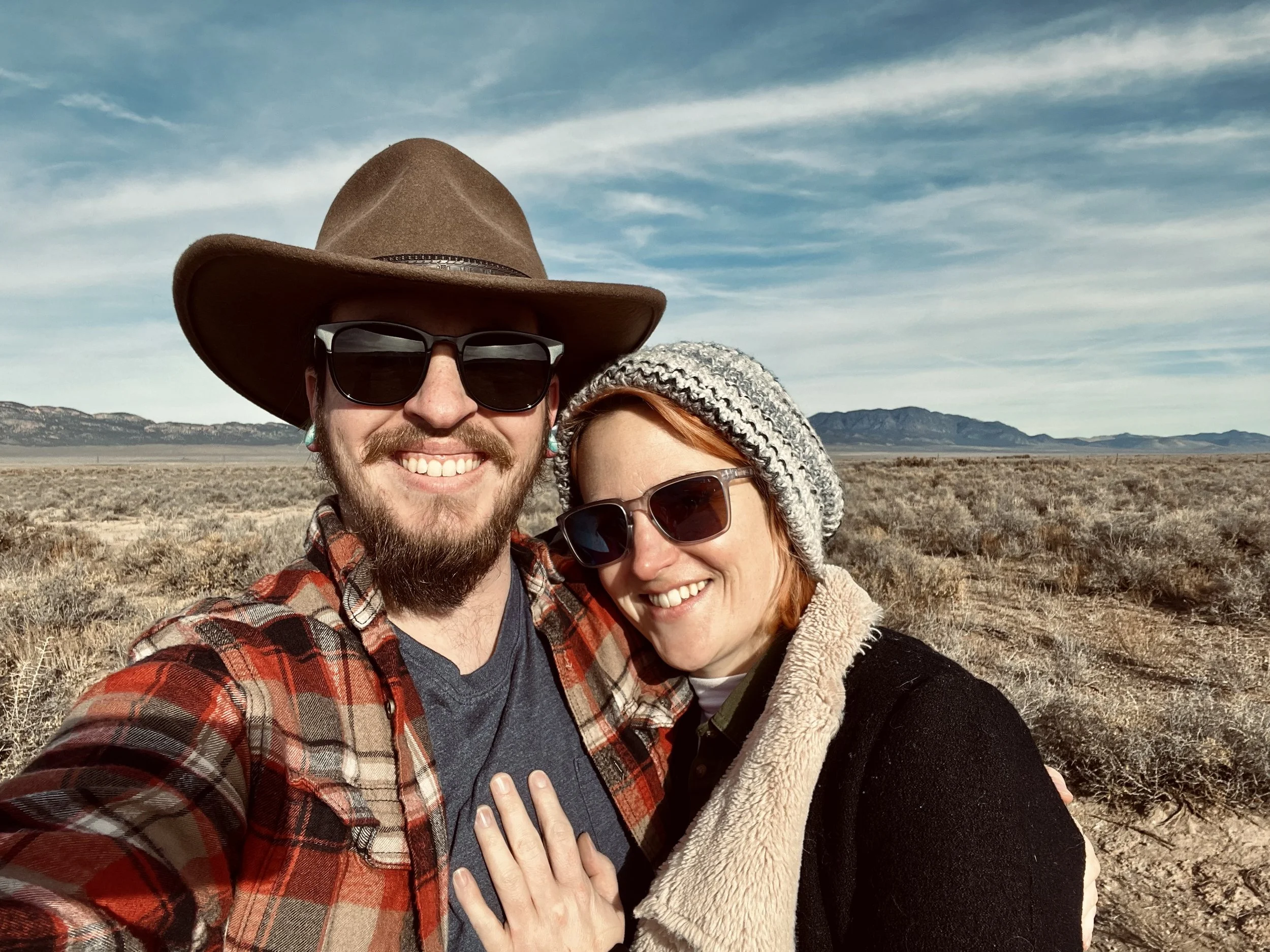 A smiling couple takes a selfie in a desert landscape with mountains in the background. Adam, is wearing a wide-brimmed hat, sunglasses, and a plaid shirt. Krys is wearing a gray knit hat, sunglasses, and a black coat with a beige collar.