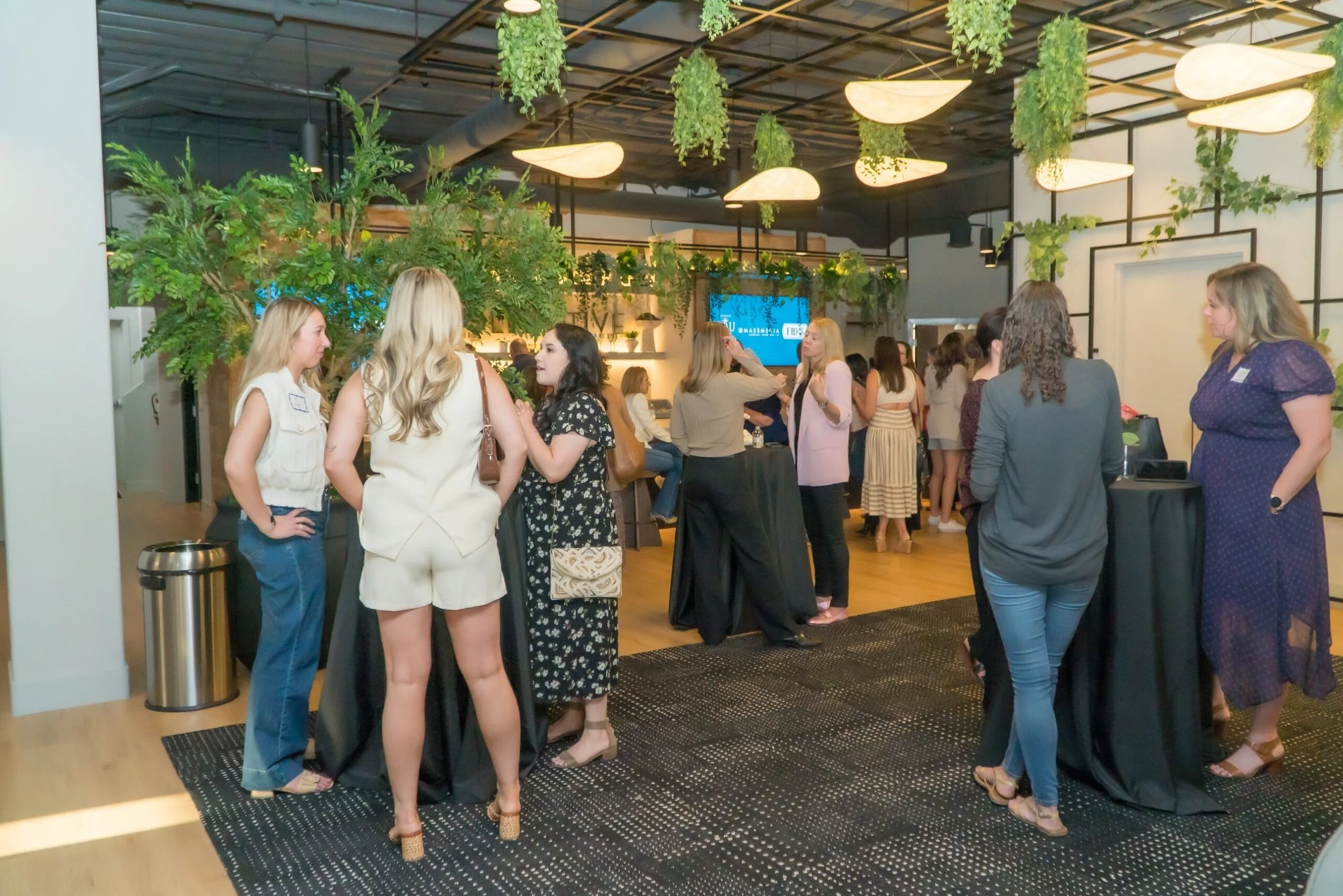 Group of women socializing at a networking event in a modern, well-lit venue with green plants and decorative lighting.