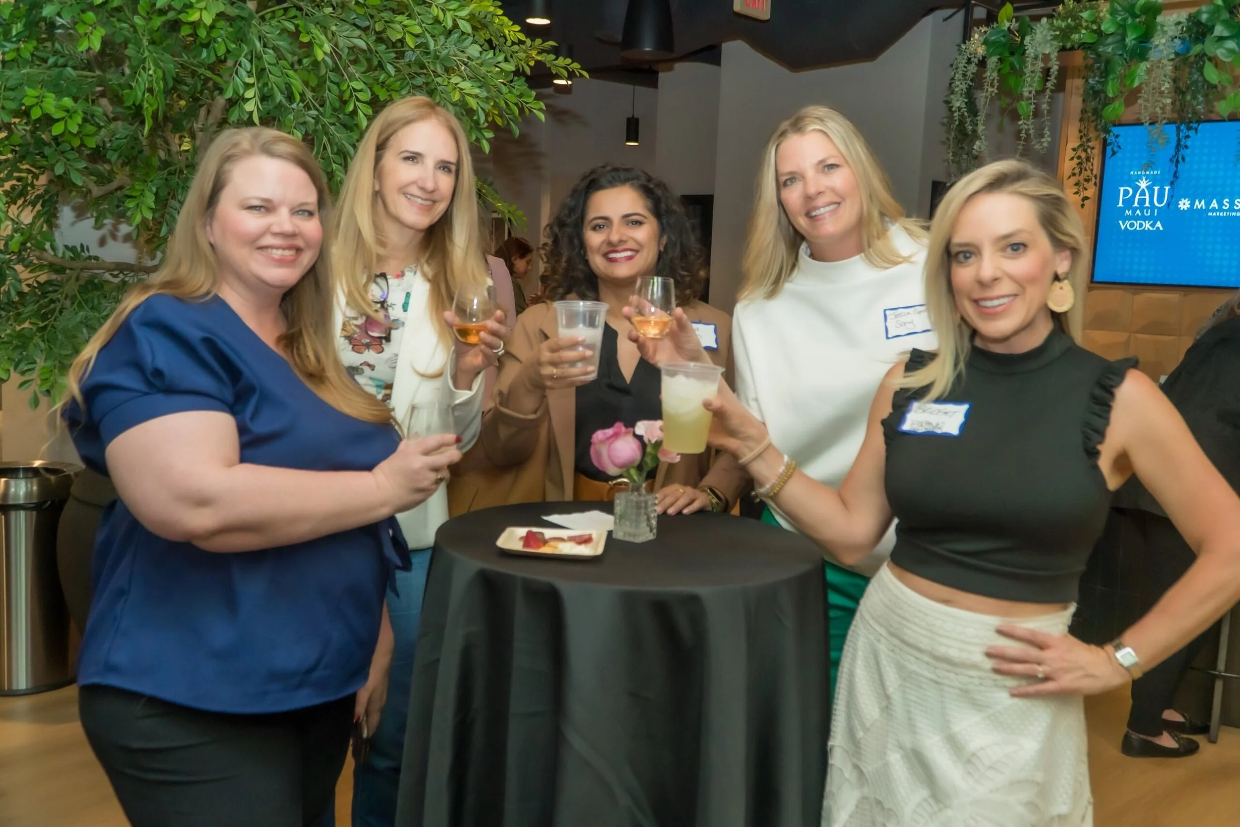 A group of six women gathered around a round table at a social event, holding drinks and smiling at the camera.