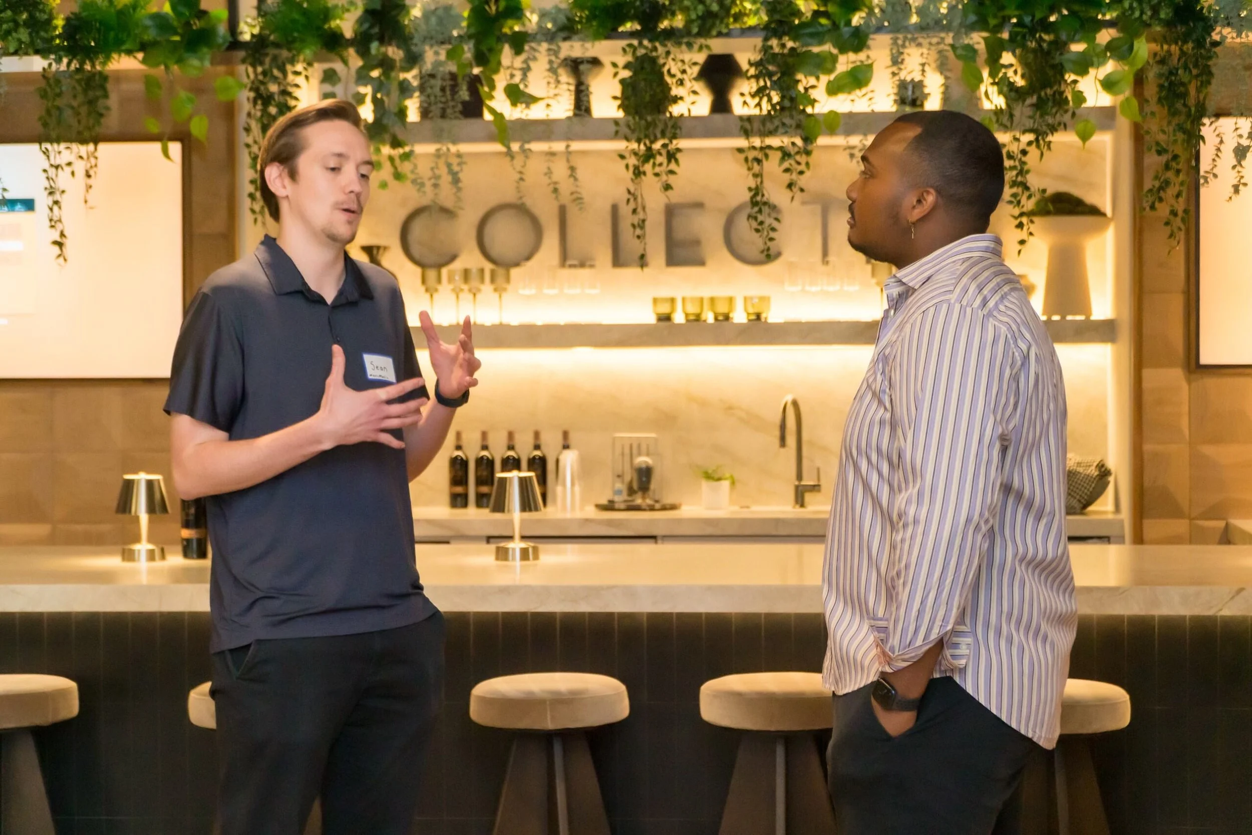 Two men having a conversation in a modern bar or restaurant with a marble counter and bar stools, greenery hanging from the ceiling, and lit shelving in the background.
