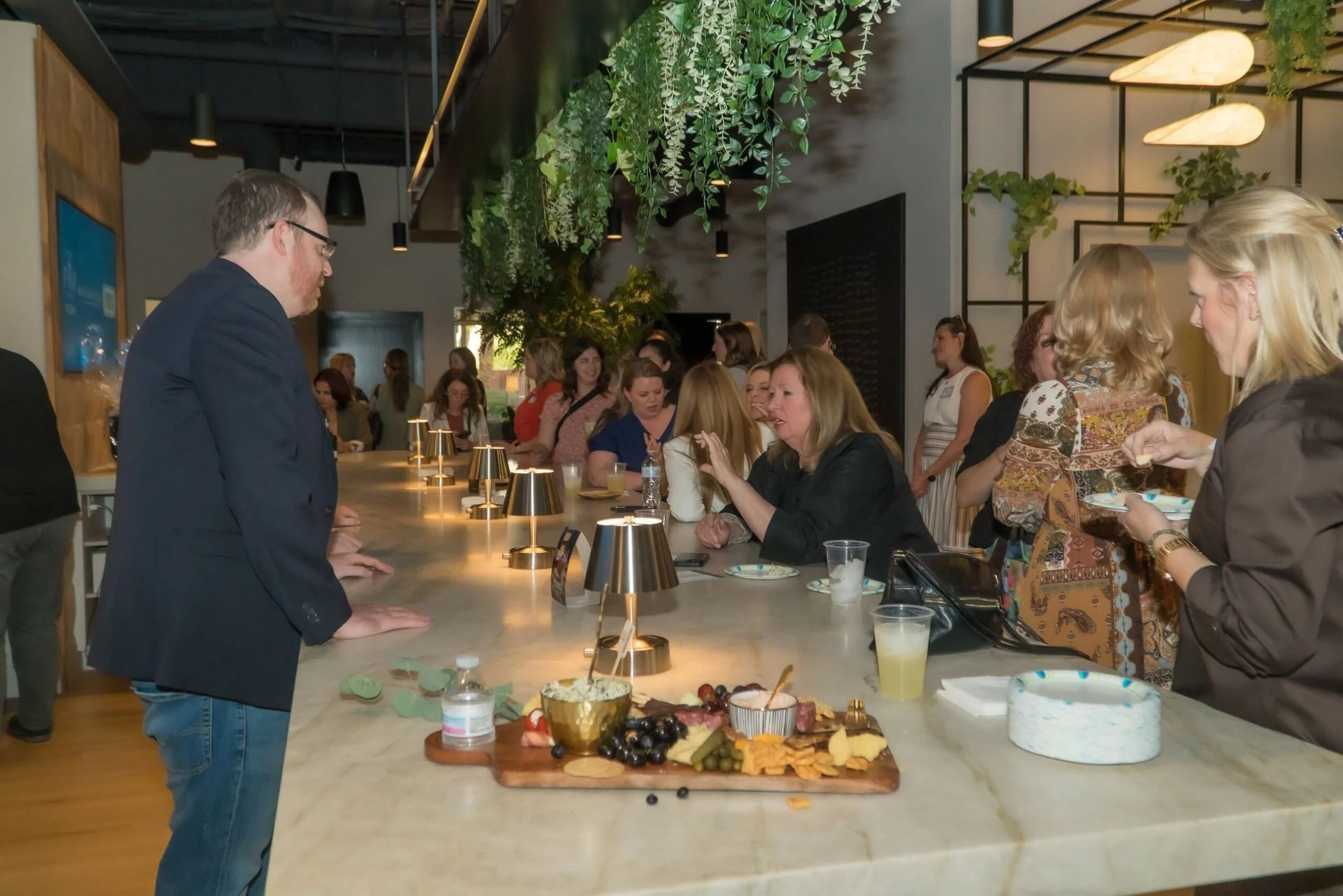 People gathered at a bar counter enjoying food and drinks, with some engaging in conversation, in a modern, well-lit venue with green hanging plants and contemporary decor.