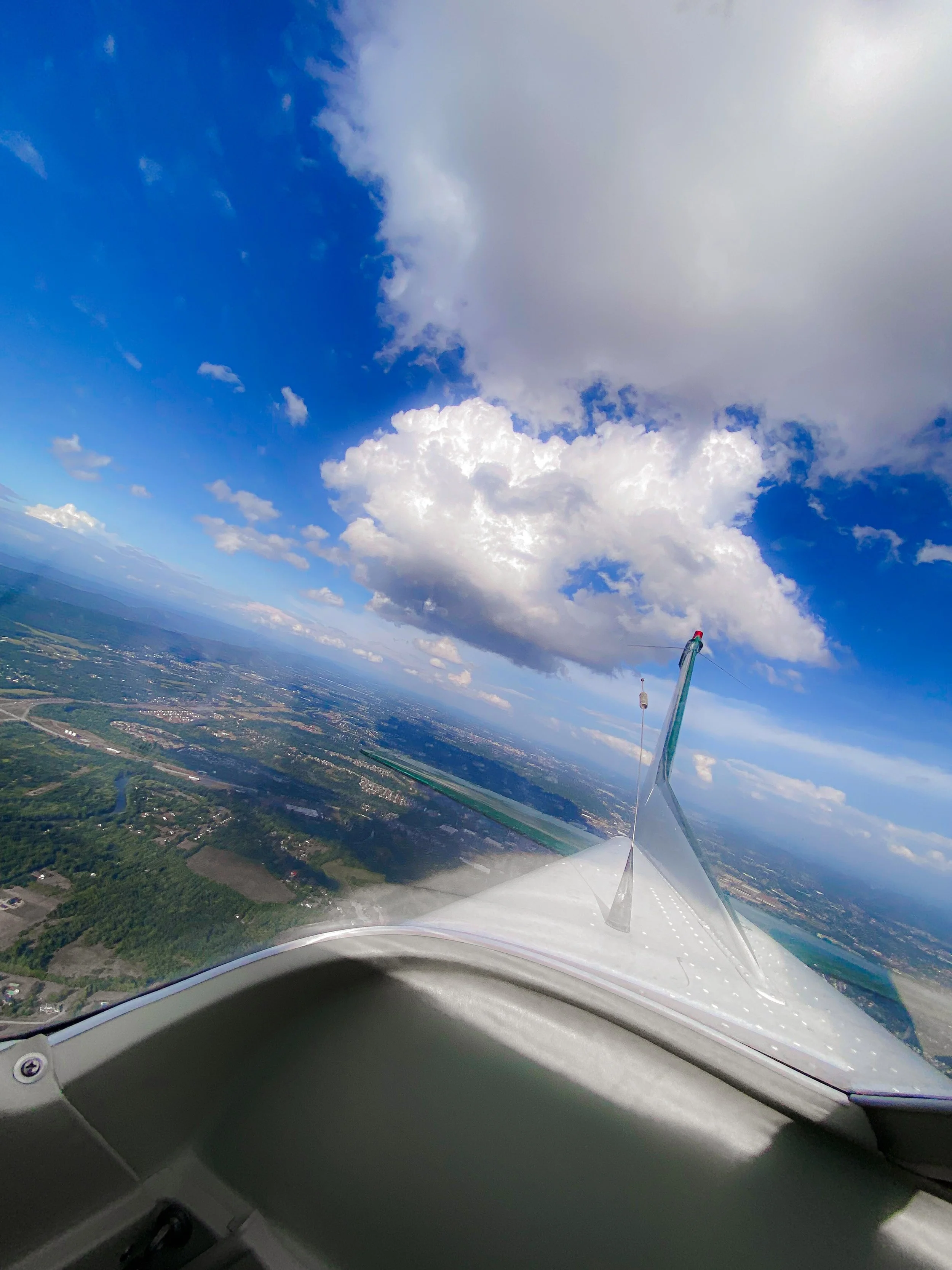 View from the cockpit of a small aircraft flying high above a landscape of fields and rivers, with partly cloudy sky overhead.
