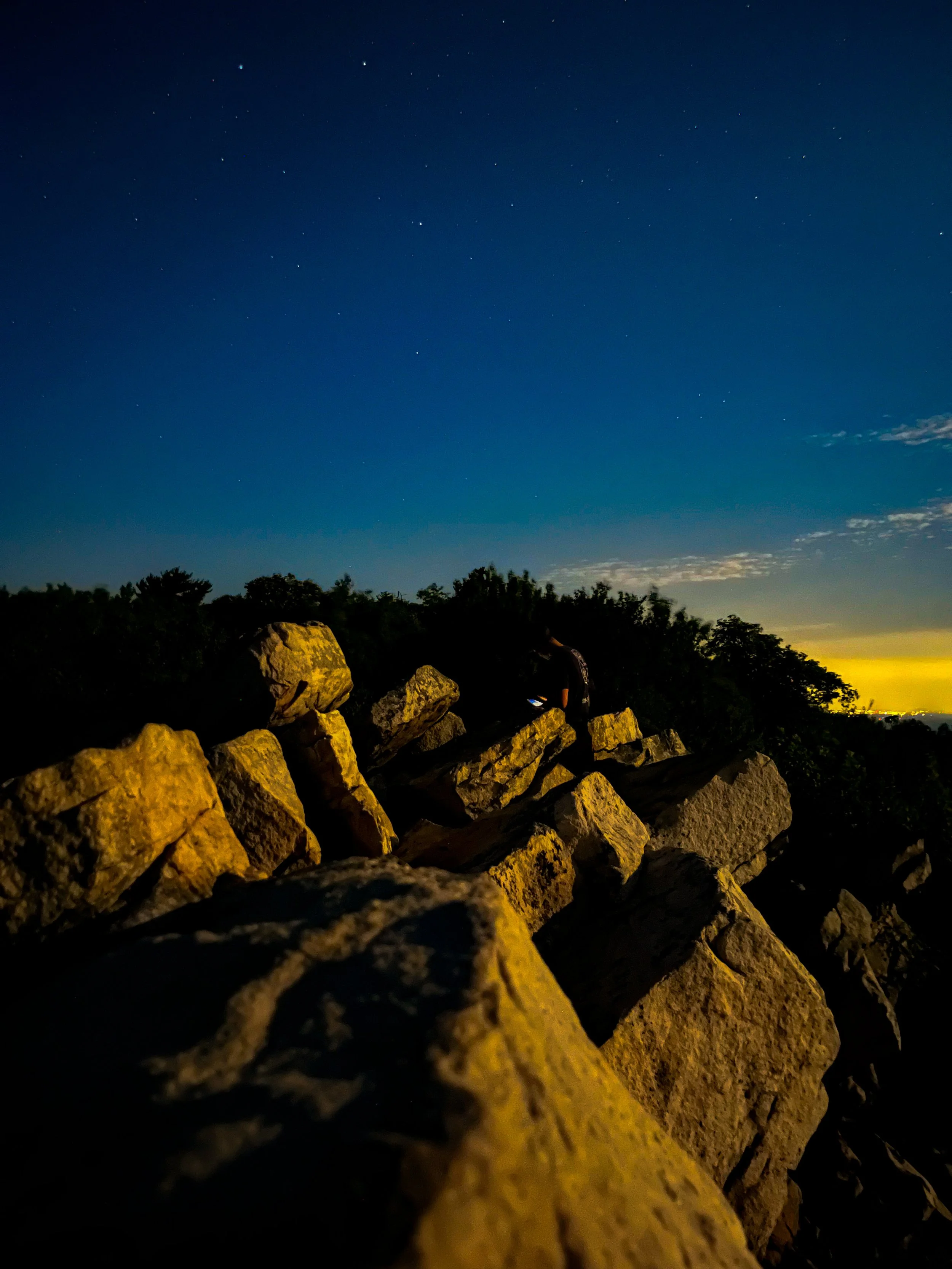 Nighttime scene with a person using a phone on a rocky ledge, starry sky above, and city lights on the horizon.